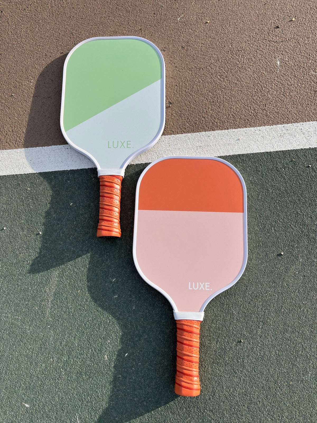 Two pickleball paddles on a tennis court. One paddle is green and white, and the other is orange and pink. Both paddles have orange grips and the word "LUXE" printed on them.