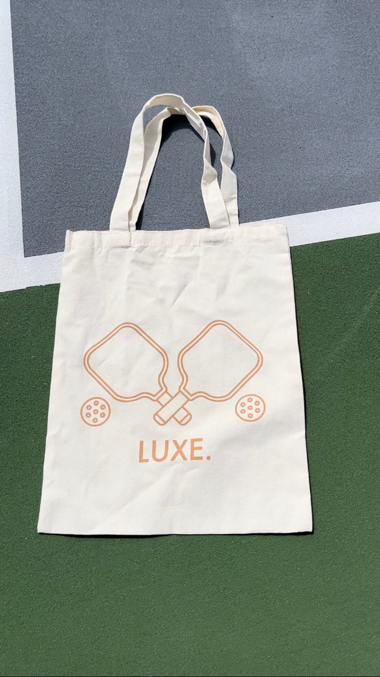 Beige tote bag with pickleball paddle and ball design, resting on a green and gray surface.