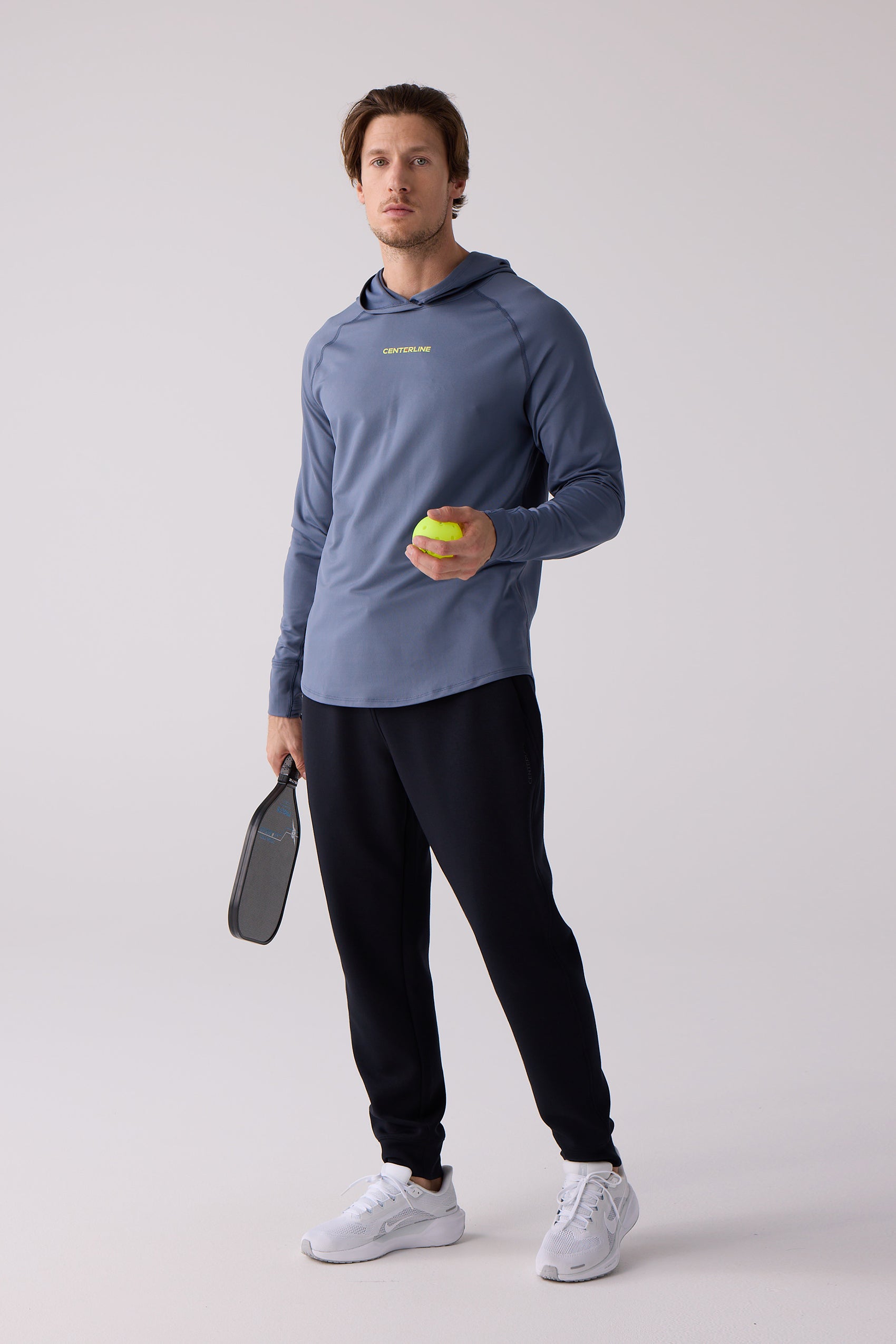 Men's blue long-sleeve layering hoodie by Centerline Athletics, featuring a hood and yellow chest logo, worn with black joggers, holding a pickleball and paddle.