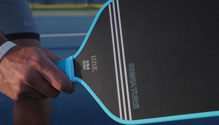 Close-up of a person holding a Luxe USA paddle with a blue edge on a tennis court.