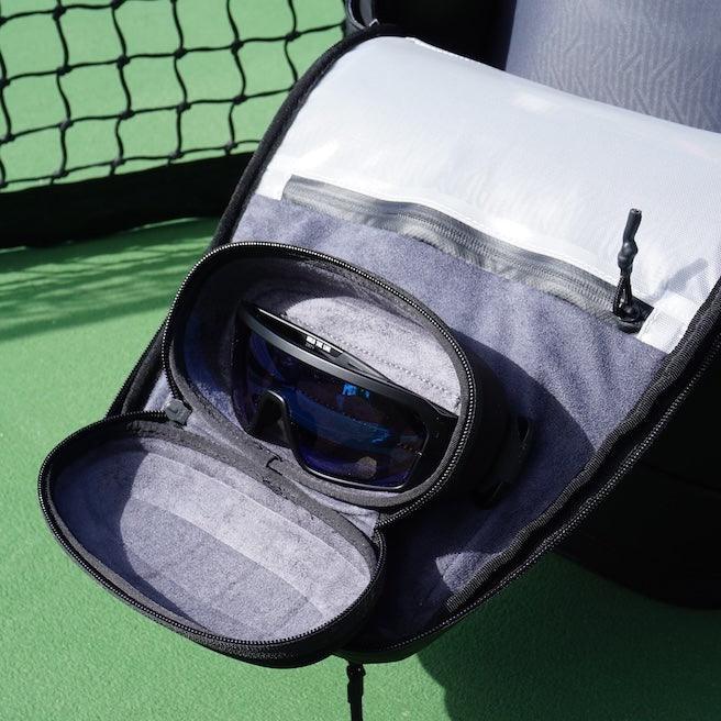 Open dark gray Magnetic Eyewear Case by FORWRD with blue-lensed sunglasses inside, on a green tennis court.