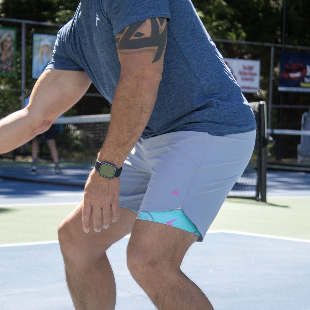 A man in a blue t-shirt and gray shorts with a blue and pink inner layer on a blue and green pickleball court.
