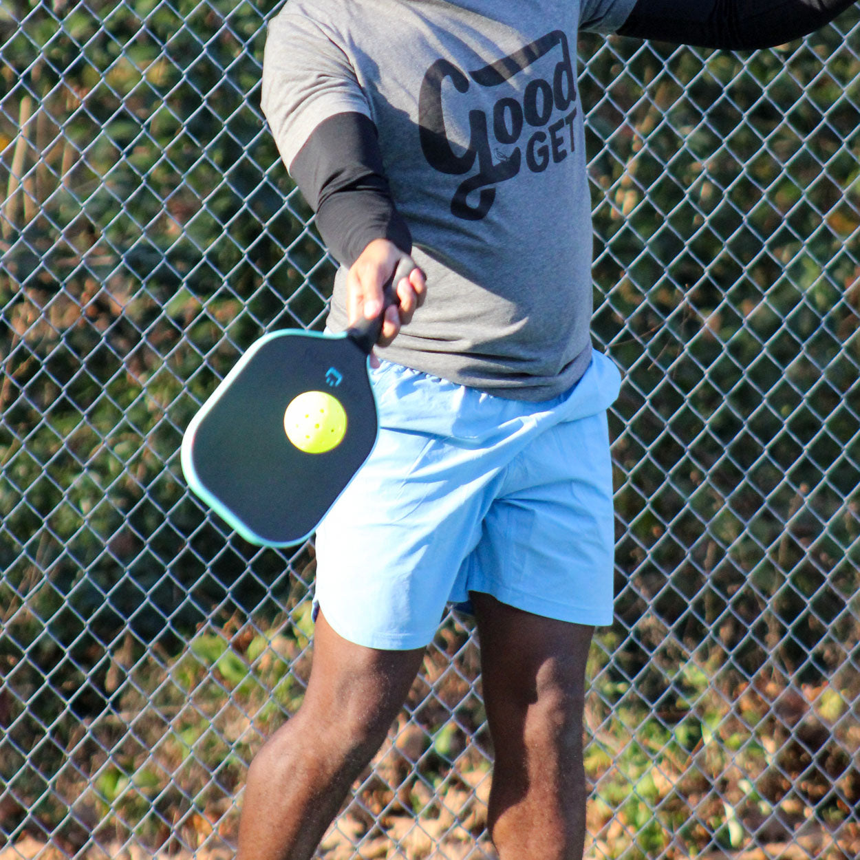 A man holding a pickleball paddle in front of a chain-link fence. He wears a grey "Good Get" t-shirt, black arm sleeves, and light blue shorts.