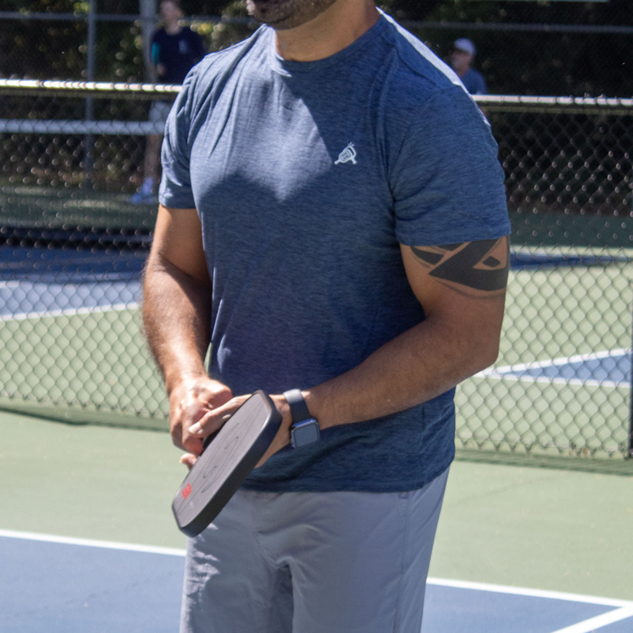Man in blue shirt with tattoo holding a black pickleball paddle.