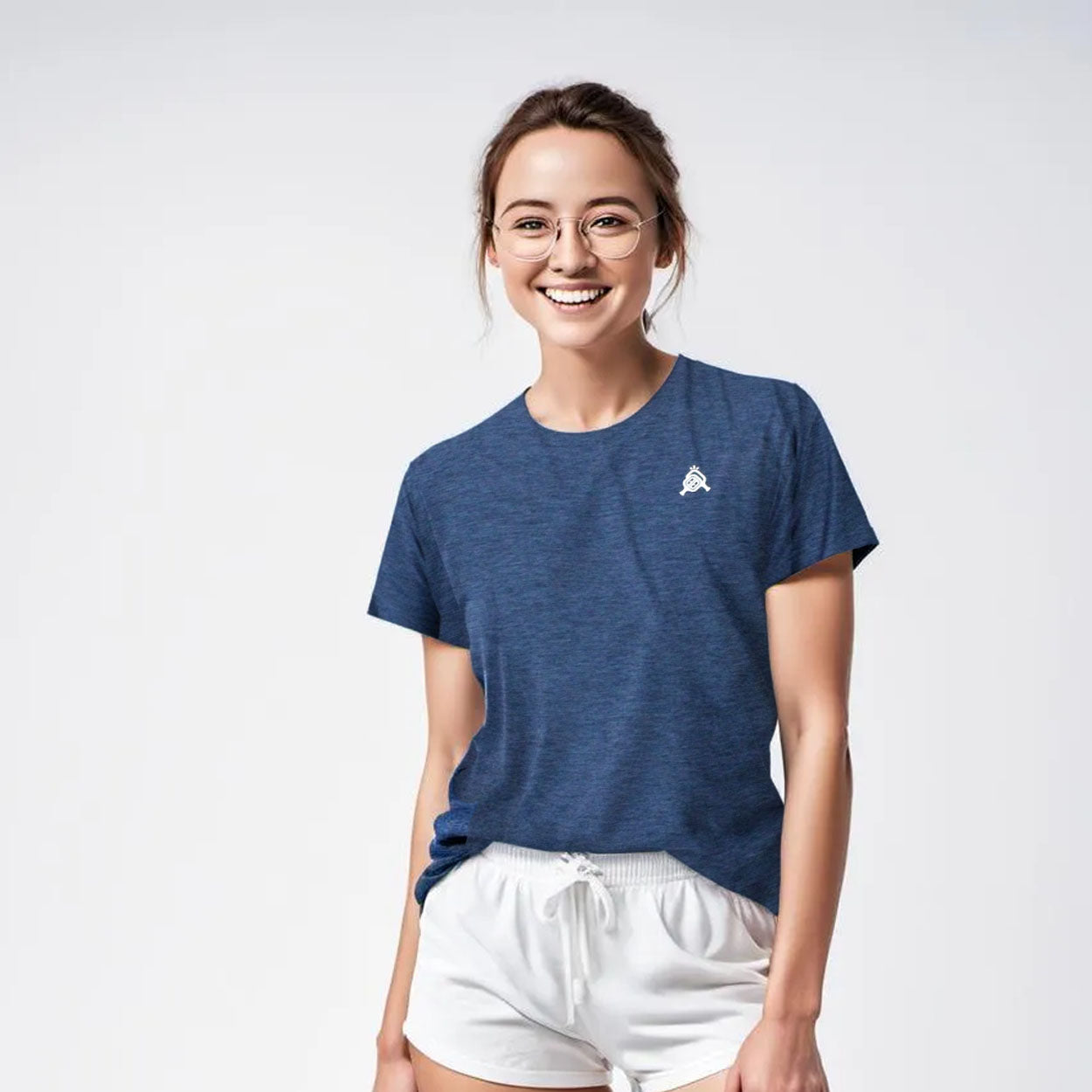 A woman in glasses, wearing a blue t-shirt with a logo and white shorts, smiles in a studio.