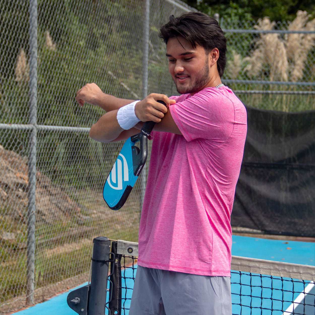 Man in pink shirt and grey shorts stretching with a blue paddle on a blue tennis court.