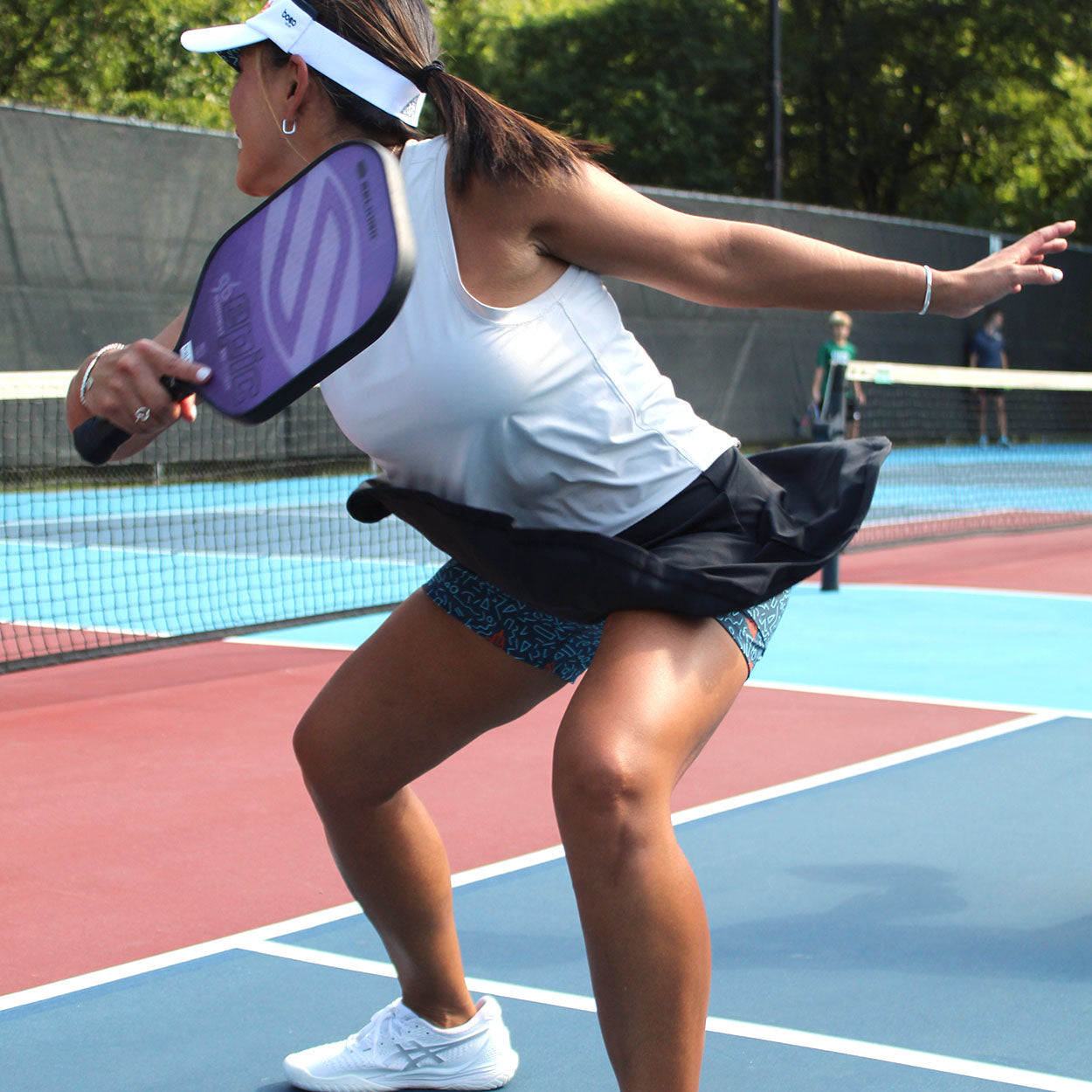 Woman wearing a white tank top, black skirt, white visor, and white tennis shoes holding a purple pickleball paddle on a blue and red court.