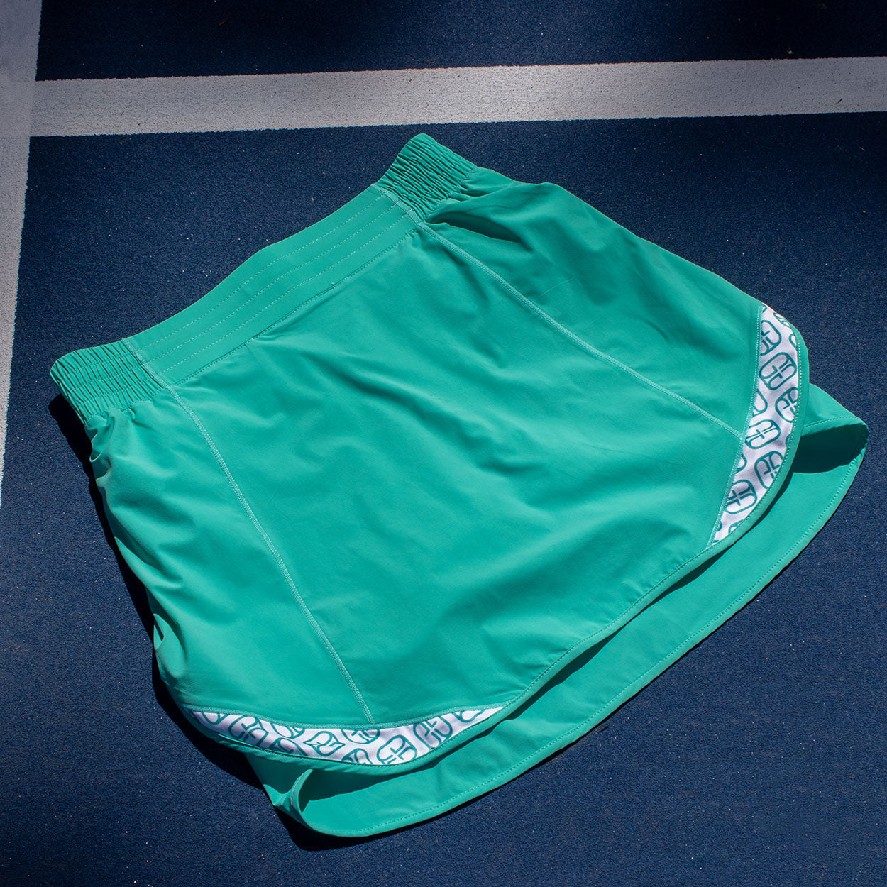 Teal pickleball skirt by Good Get with white and teal logo patterns on side inserts, on a blue court.
