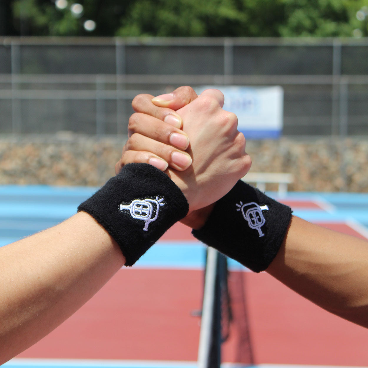 Two people shaking hands wearing black wristbands with a white logo on a tennis court.