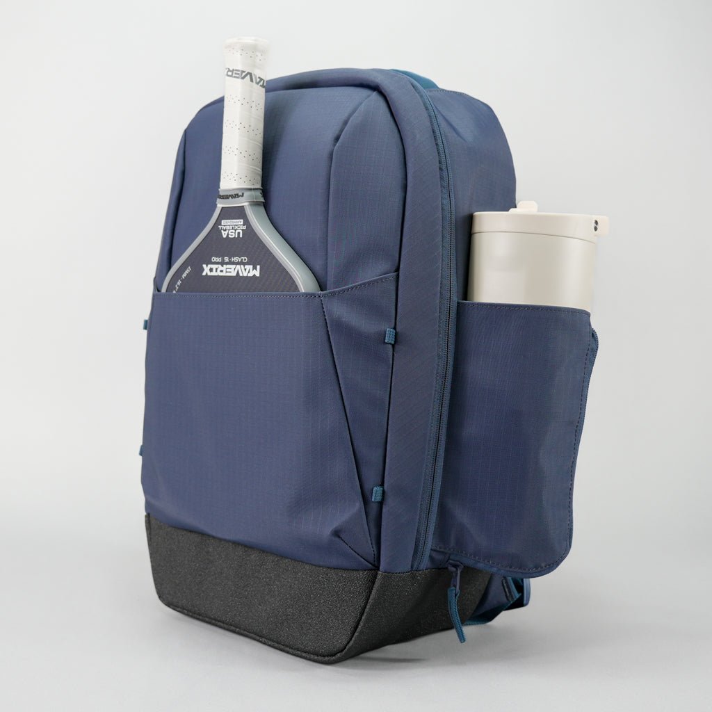 Navy blue textured pickleball backpack by FORWRD, with a black base. It holds a white-gripped pickleball paddle and an off-white tumbler in its side pockets.