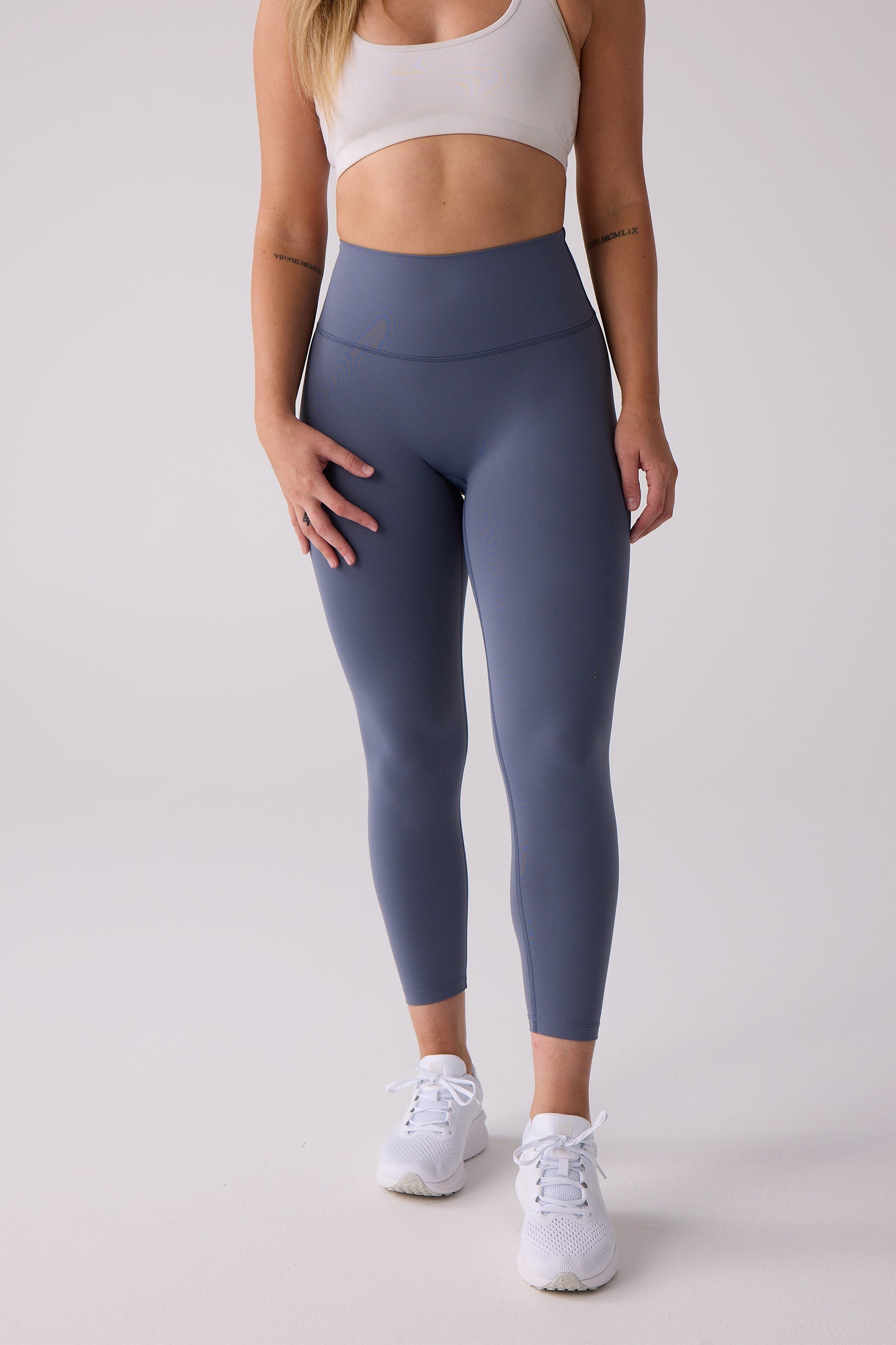 Slate blue 7/8 legging by Centerline Athletics, smooth material, paired with a white sports bra and sneakers.
