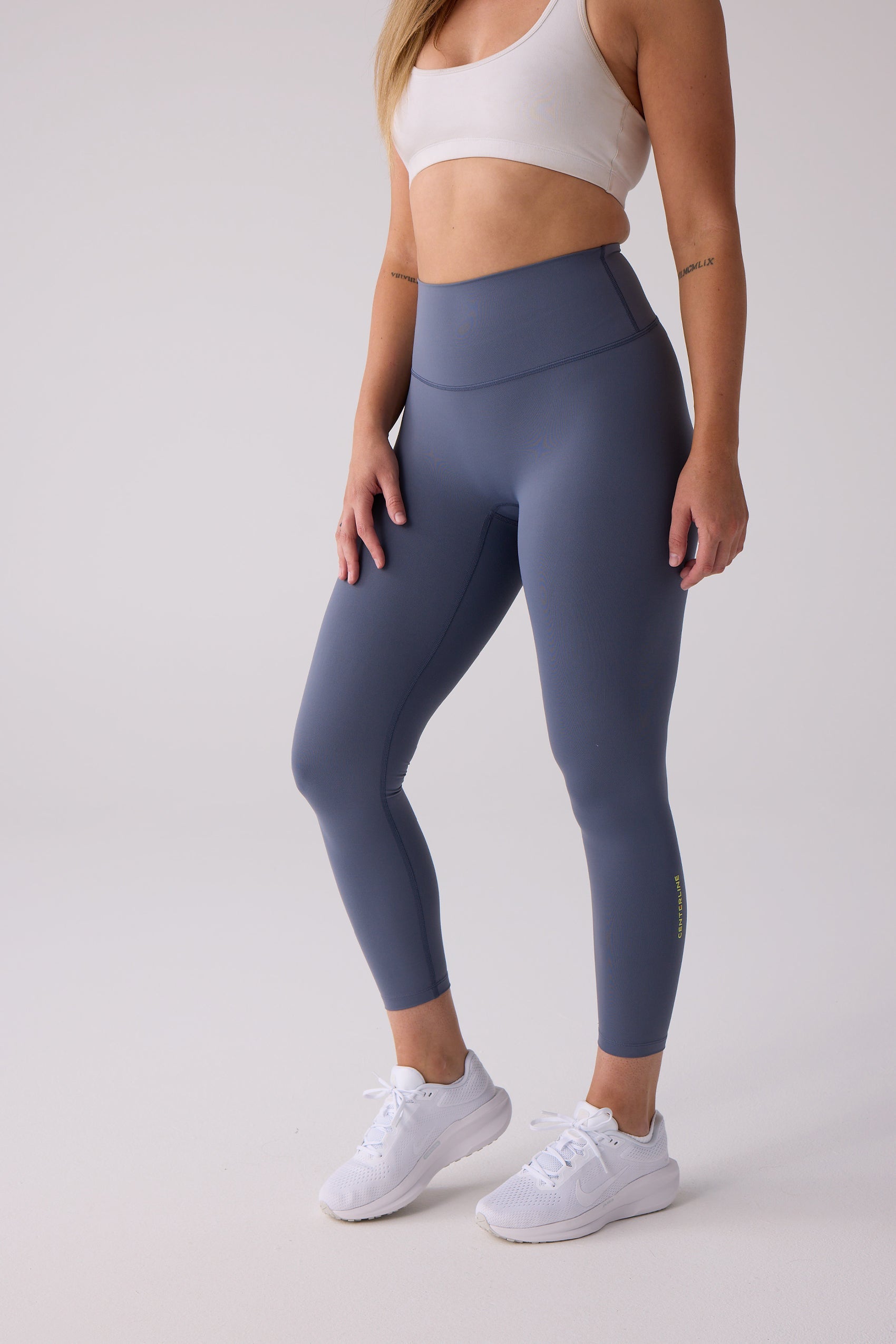 Dark blue 7/8 leggings from Centerline Athletics with vertical yellow 'CENTERLINE' text on the lower leg, paired with a white sports bra and sneakers.