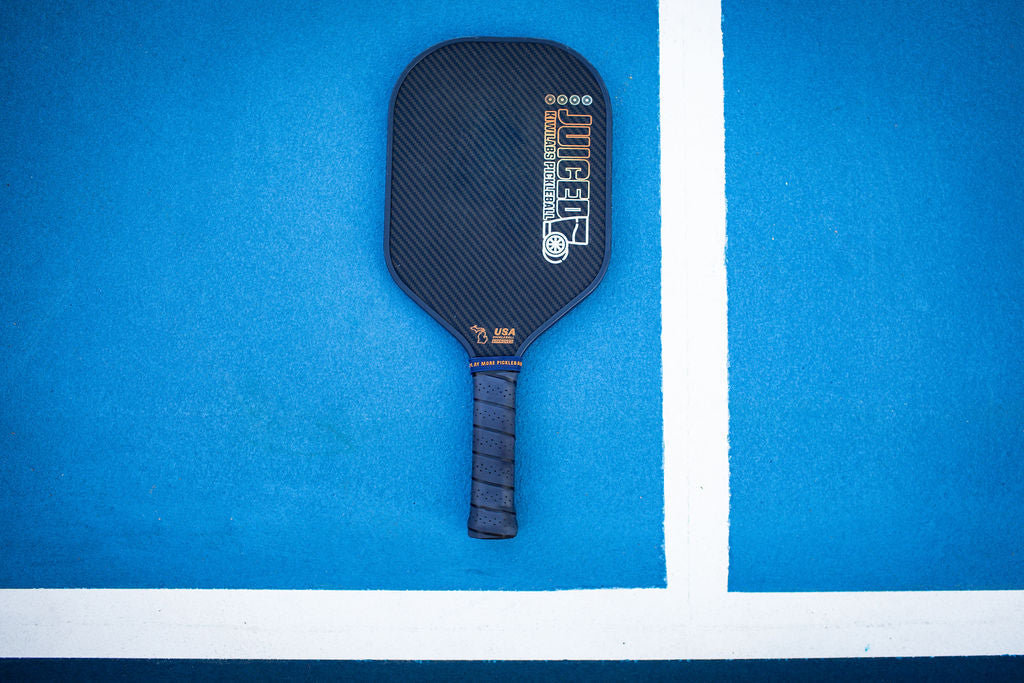 Close-up of a black Juiced KiwiLabs Pickleball paddle on a blue court with white lines.