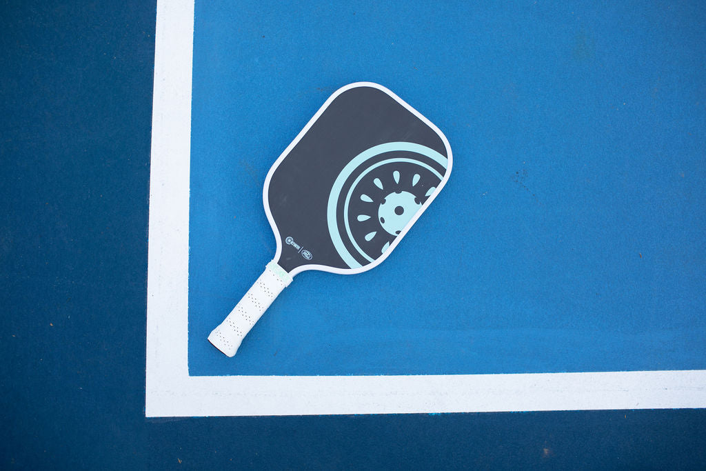 Overhead shot of a blue and gray pickleball paddle on a blue pickleball court with white lines.