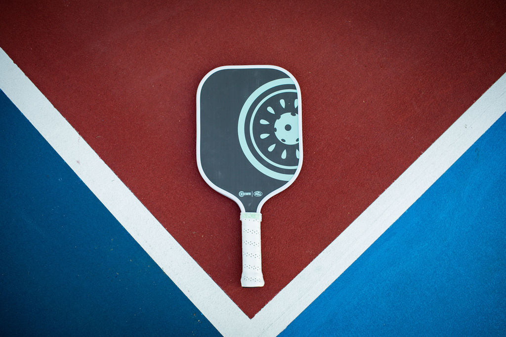 Pickleball paddle on a court with red and blue zones, divided by white lines.