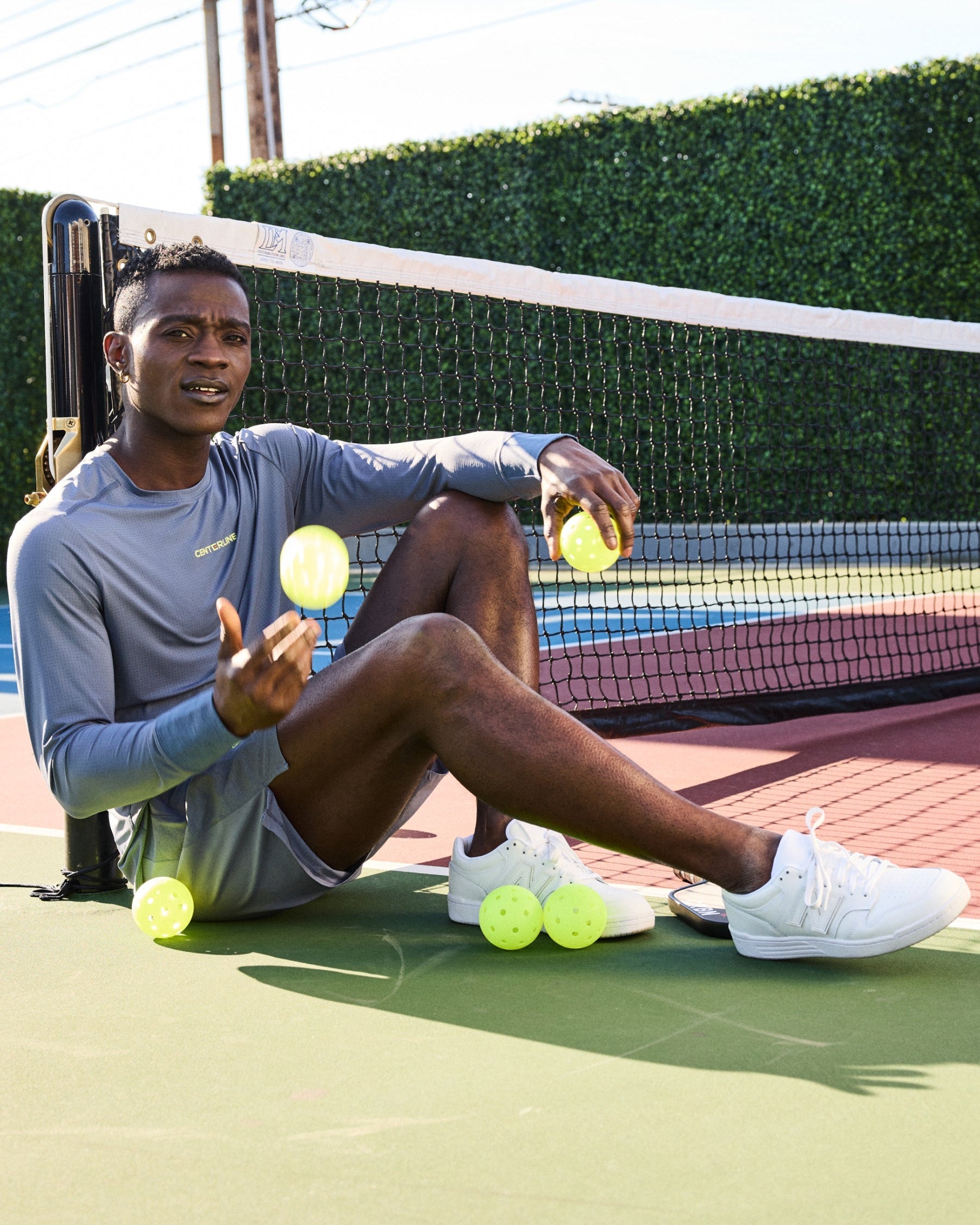 A Black man sits on a pickleball court, wearing a gray long-sleeve shirt and shorts, white tennis shoes, holding and surrounded by yellow pickleballs.