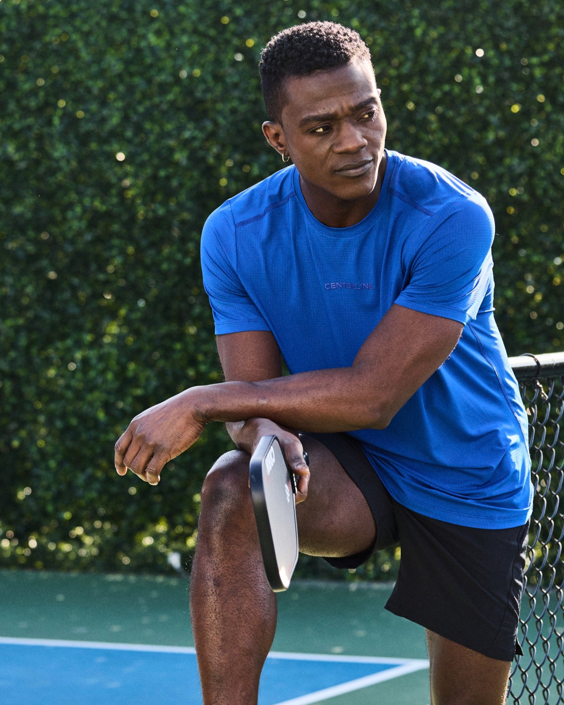 Man leaning on a fence wearing a blue athletic shirt, black shorts, and holding a pickleball paddle.
