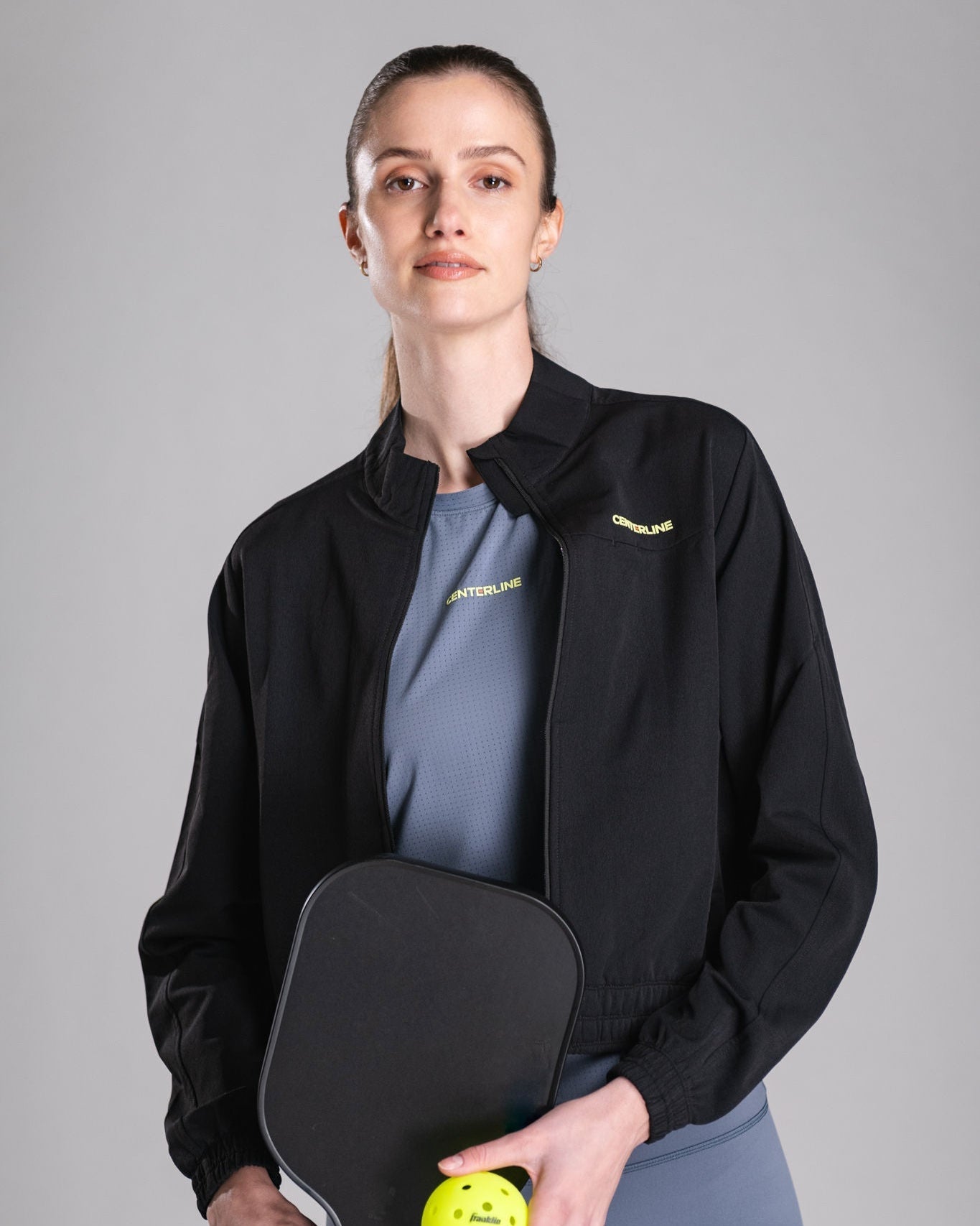 Woman holding pickleball paddle and ball, wearing black jacket and gray shirt.