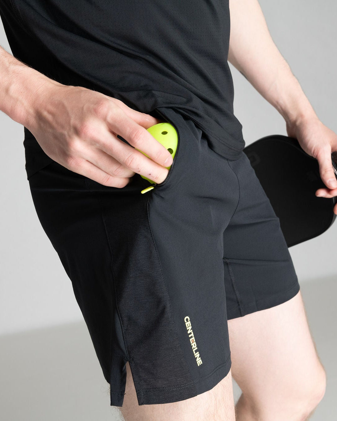 A man puts a yellow pickleball into the zippered pocket of black athletic shorts.