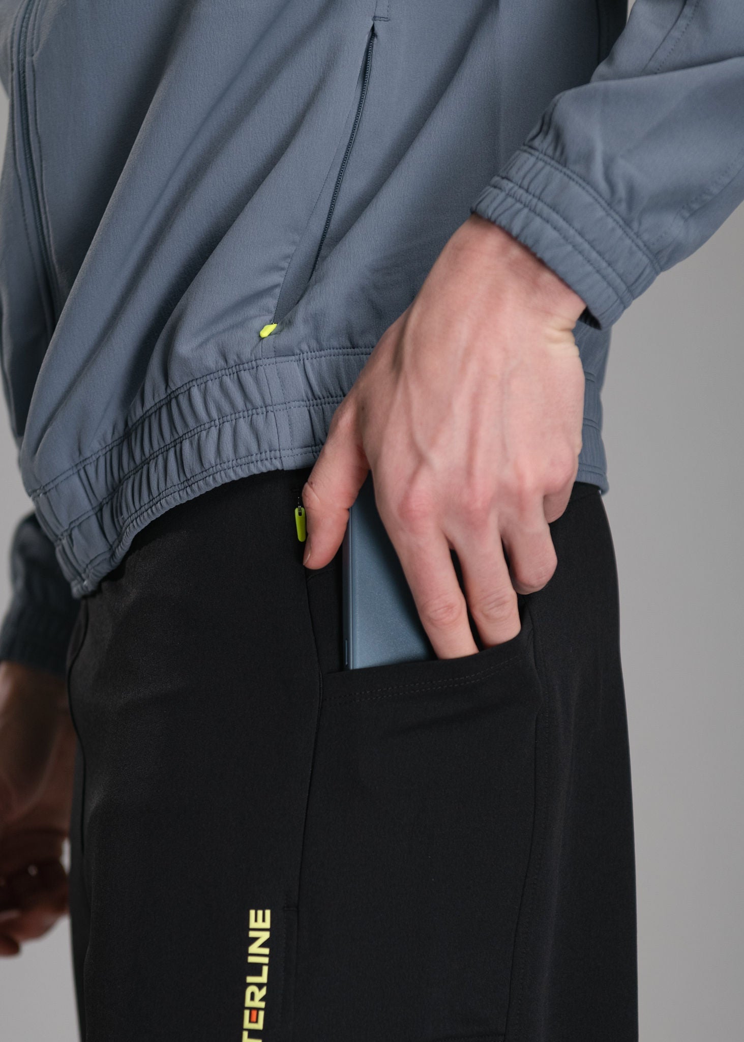 Person putting a blue cell phone in the pocket of black pants, while wearing a gray jacket.