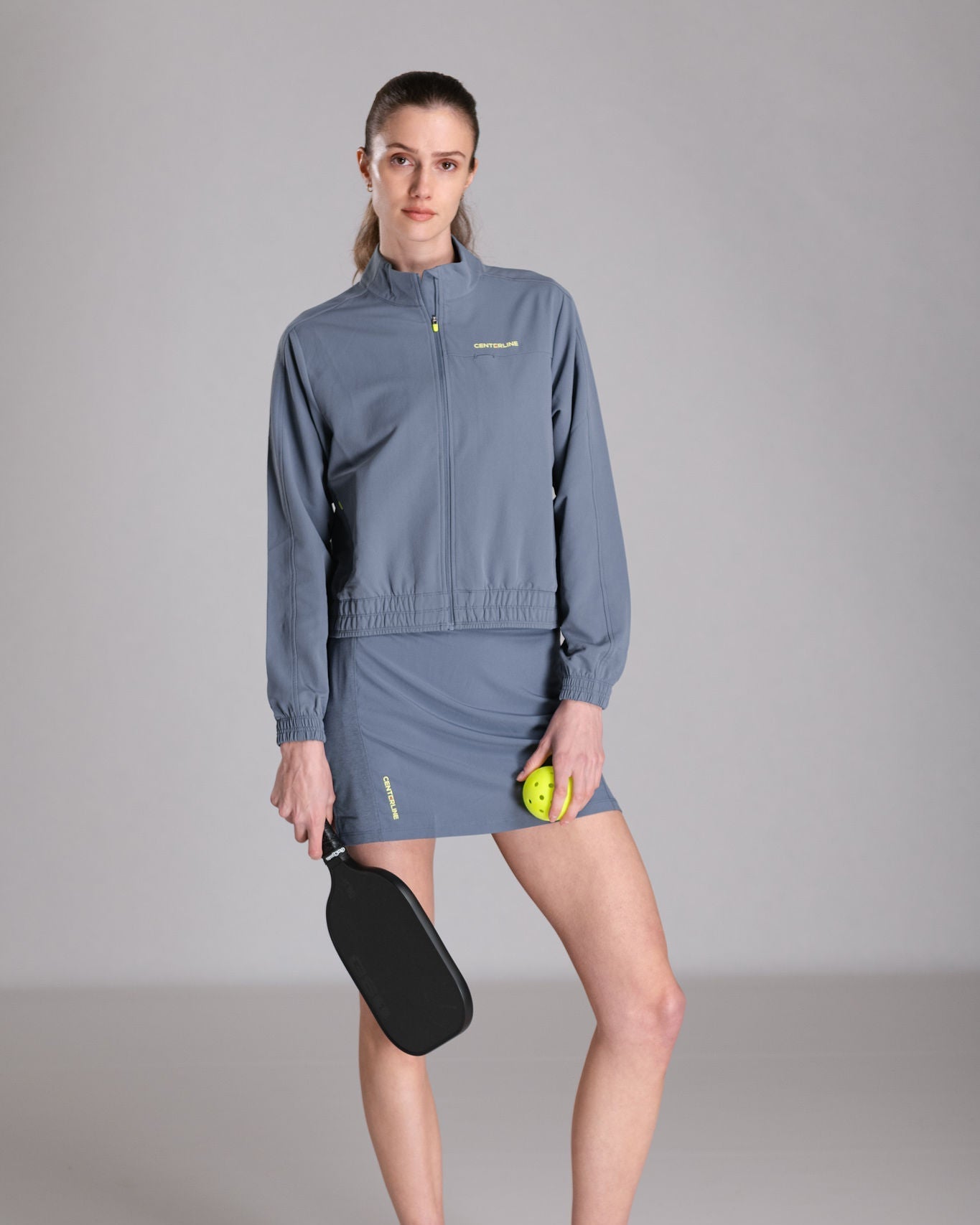 Woman in a blue matching jacket and skirt holding a pickleball paddle and ball.