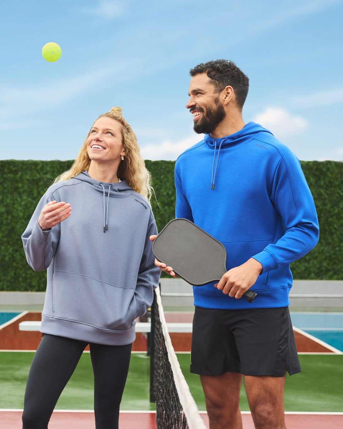 Two people wearing athletic hoodies, playing pickleball outdoors.