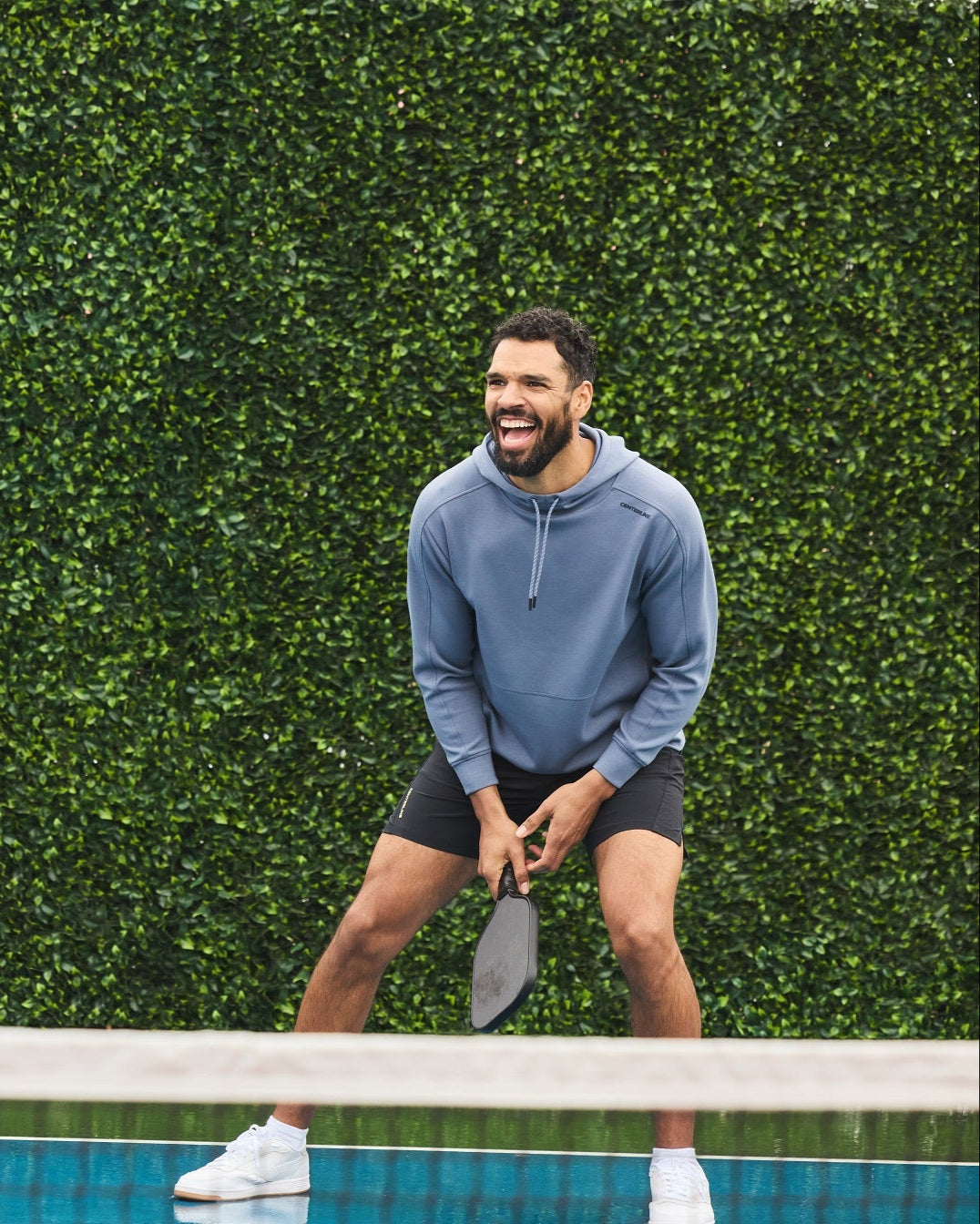 Man laughing and playing pickleball, wearing a blue hoodie and black shorts.
