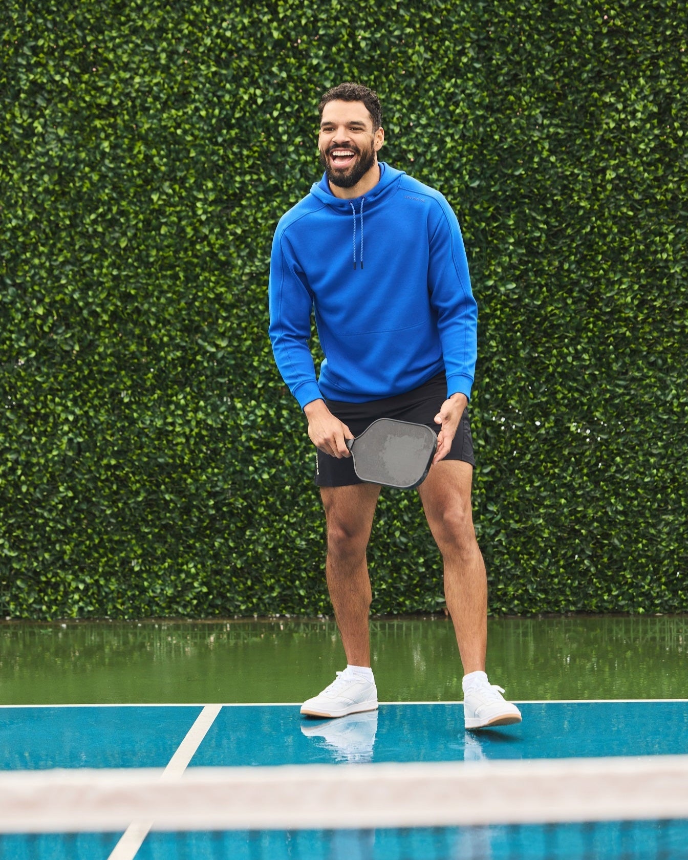 A man in a blue hoodie and black shorts plays pickleball on a blue court.
