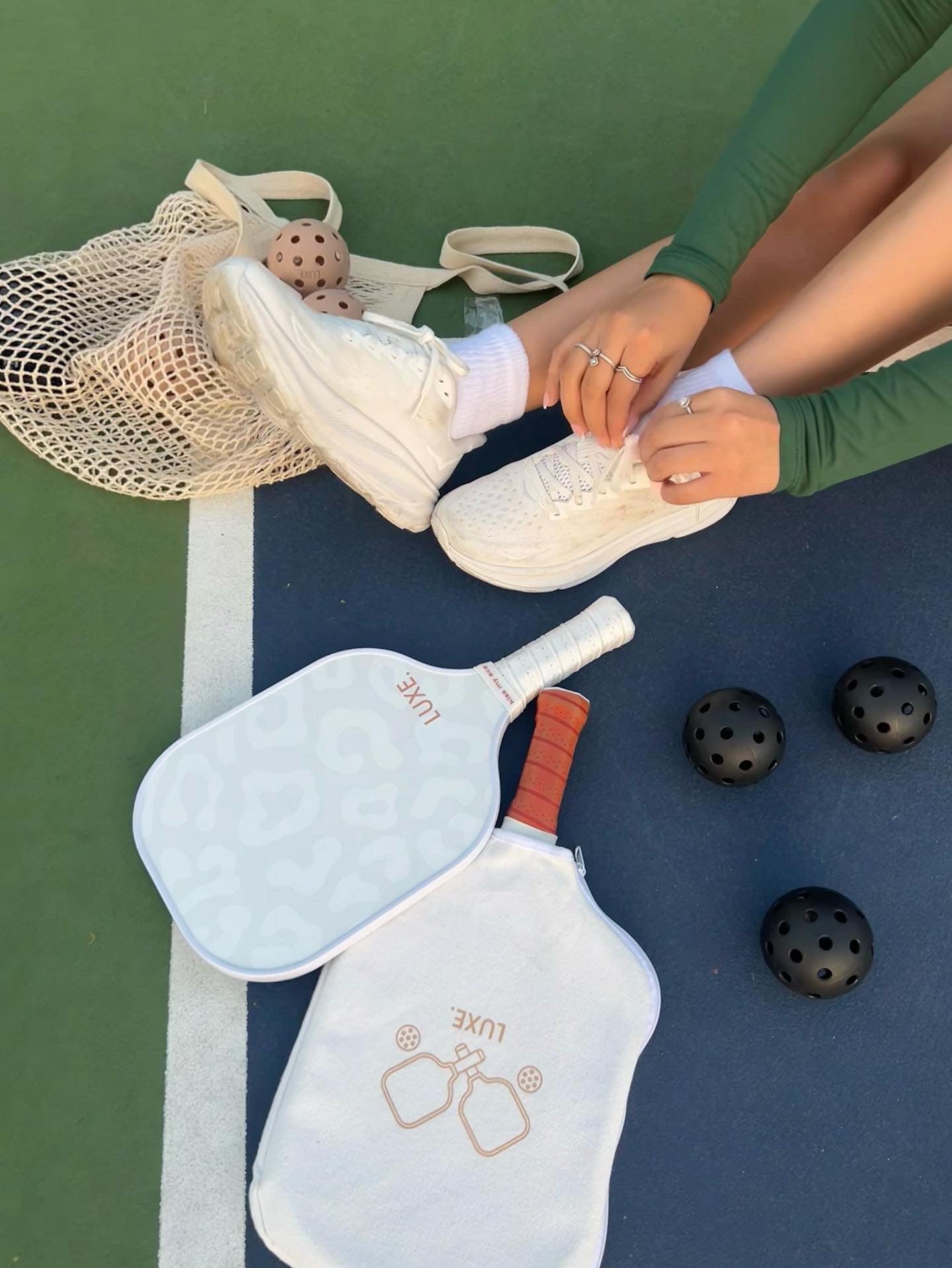 Person tying white shoes on a blue and green pickleball court with paddles and black balls.
