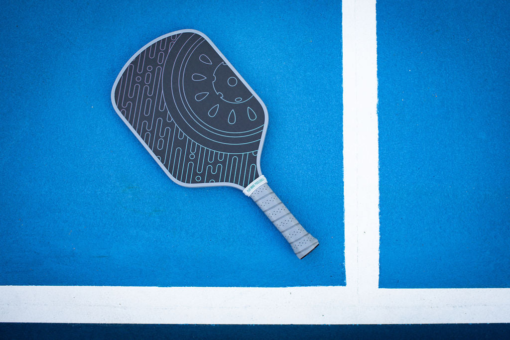 Overhead shot of a gray pickleball paddle with a watermelon graphic on a blue pickleball court with white lines.