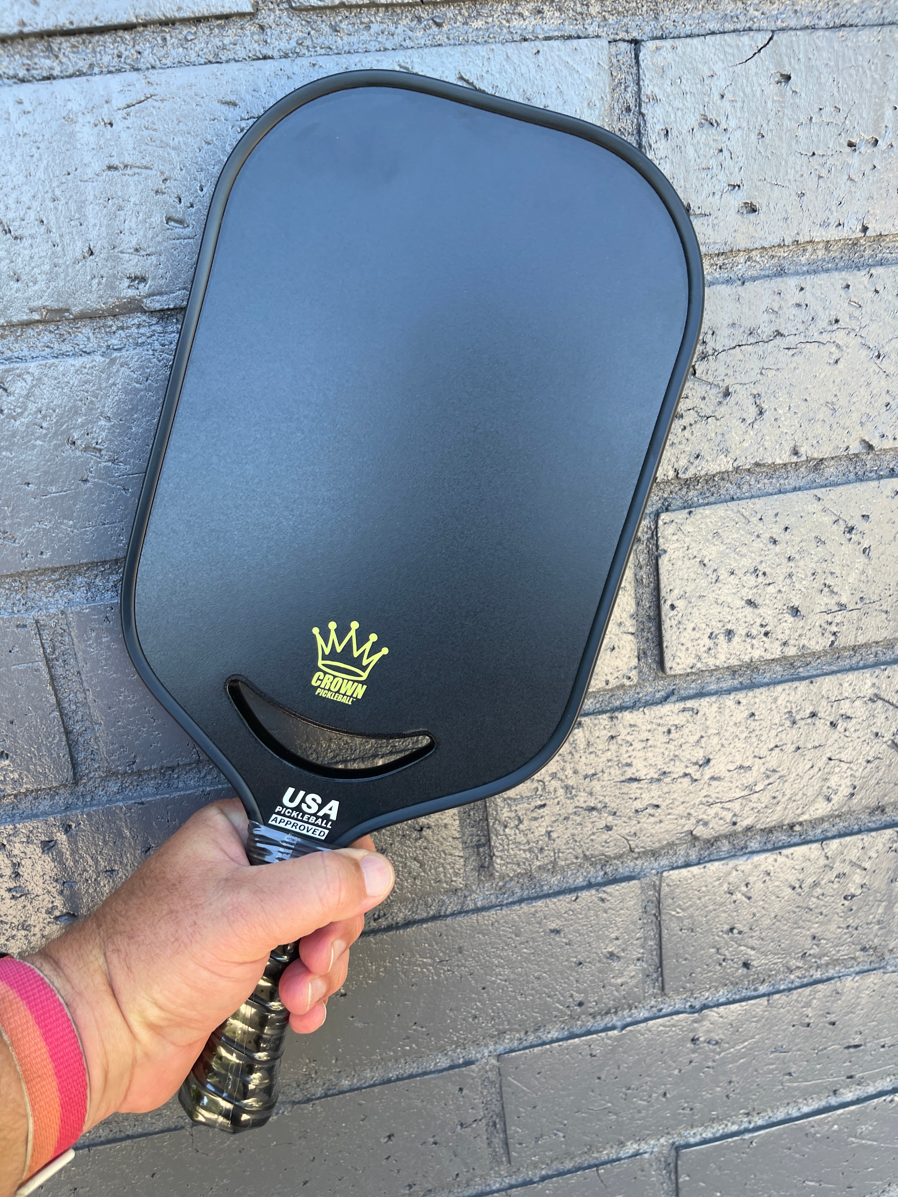 Close-up of a black Crown Pickleball paddle against a painted brick wall, being held by a hand wearing a pink and orange wristband.