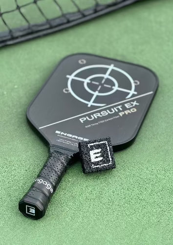 Black, square, textured pickleball paddle cleaner by EngagePickleball, with a white 'E' logo, on a court beside a black paddle.