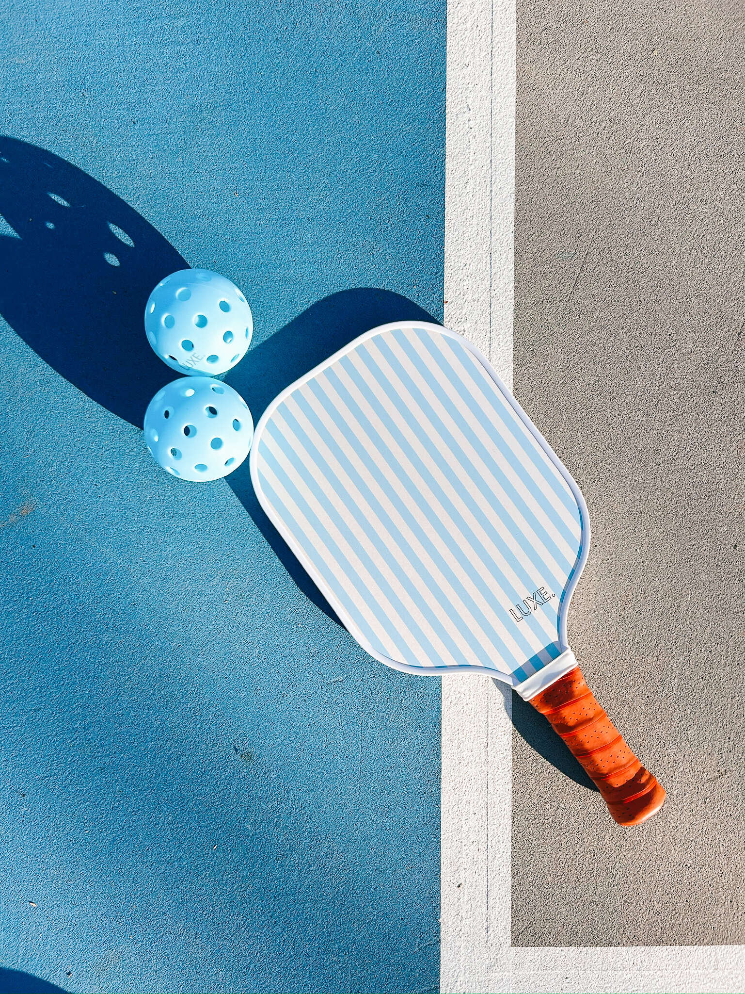 Two light blue pickleballs and a striped paddle on a blue and gray court.