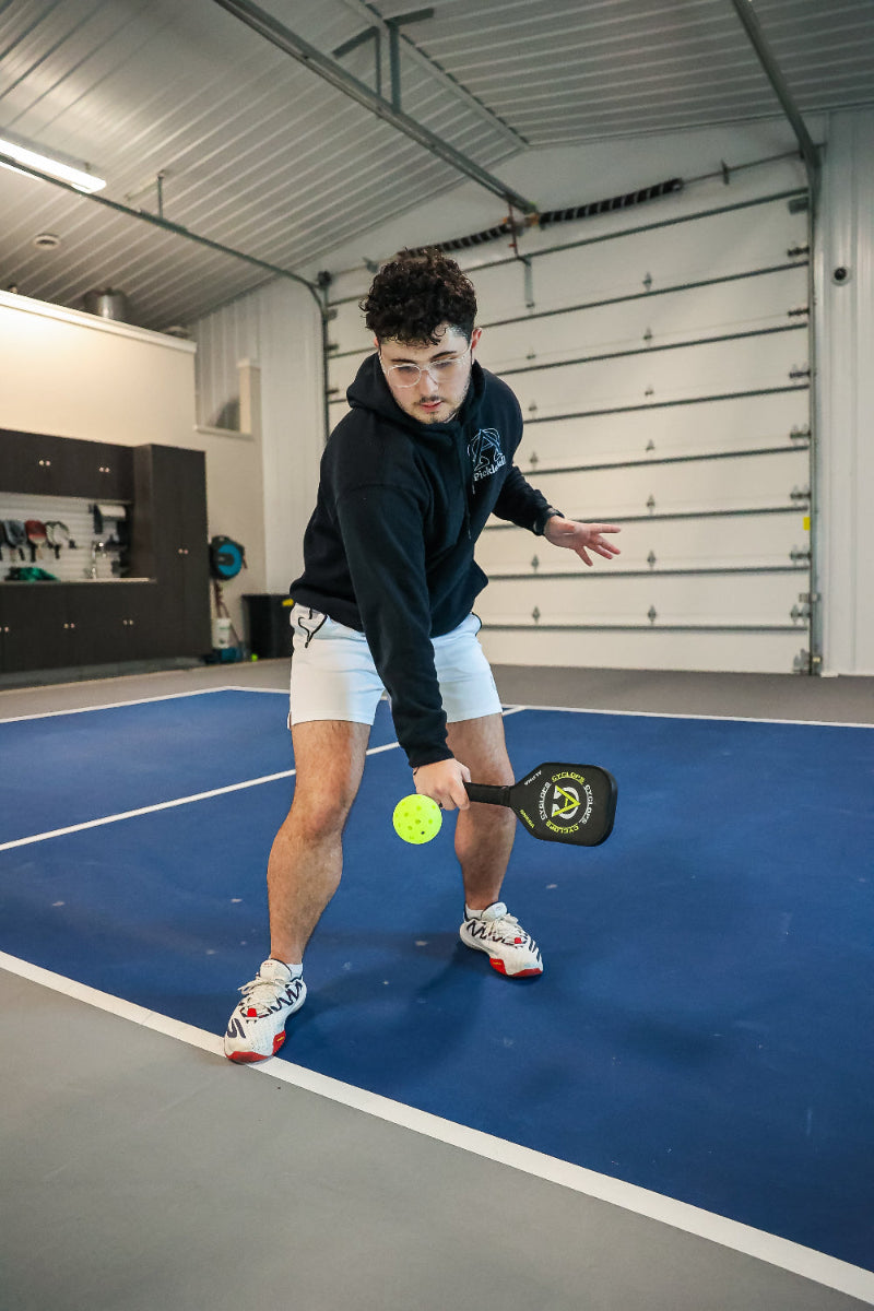 A man playing pickleball in a gym, wearing a black hoodie, white shorts, and sneakers.