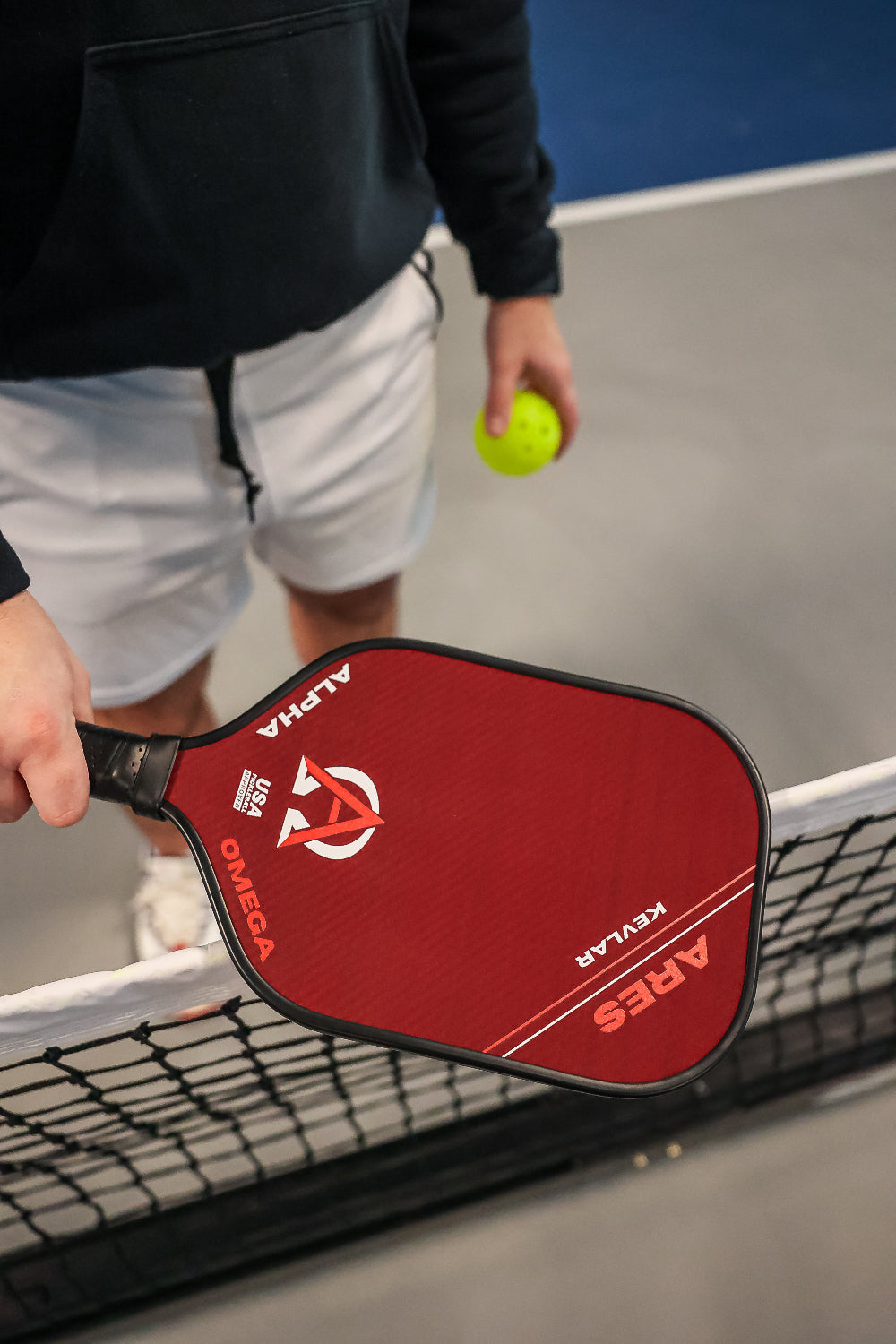 A person wearing a black hoodie and white shorts holding a pickleball and a red Ares Kevlar pickleball paddle over a net.