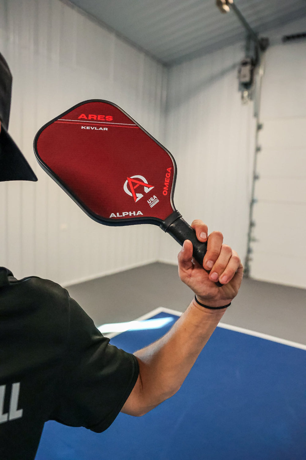 Man holding a red and black Ares Kevlar pickleball paddle indoors.