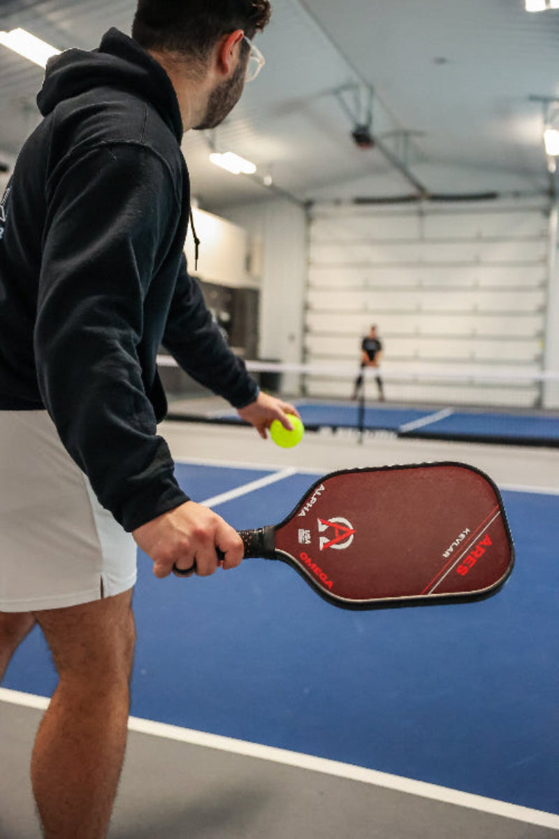 Man in black hoodie and white shorts playing pickleball, holding a red paddle and yellow ball.