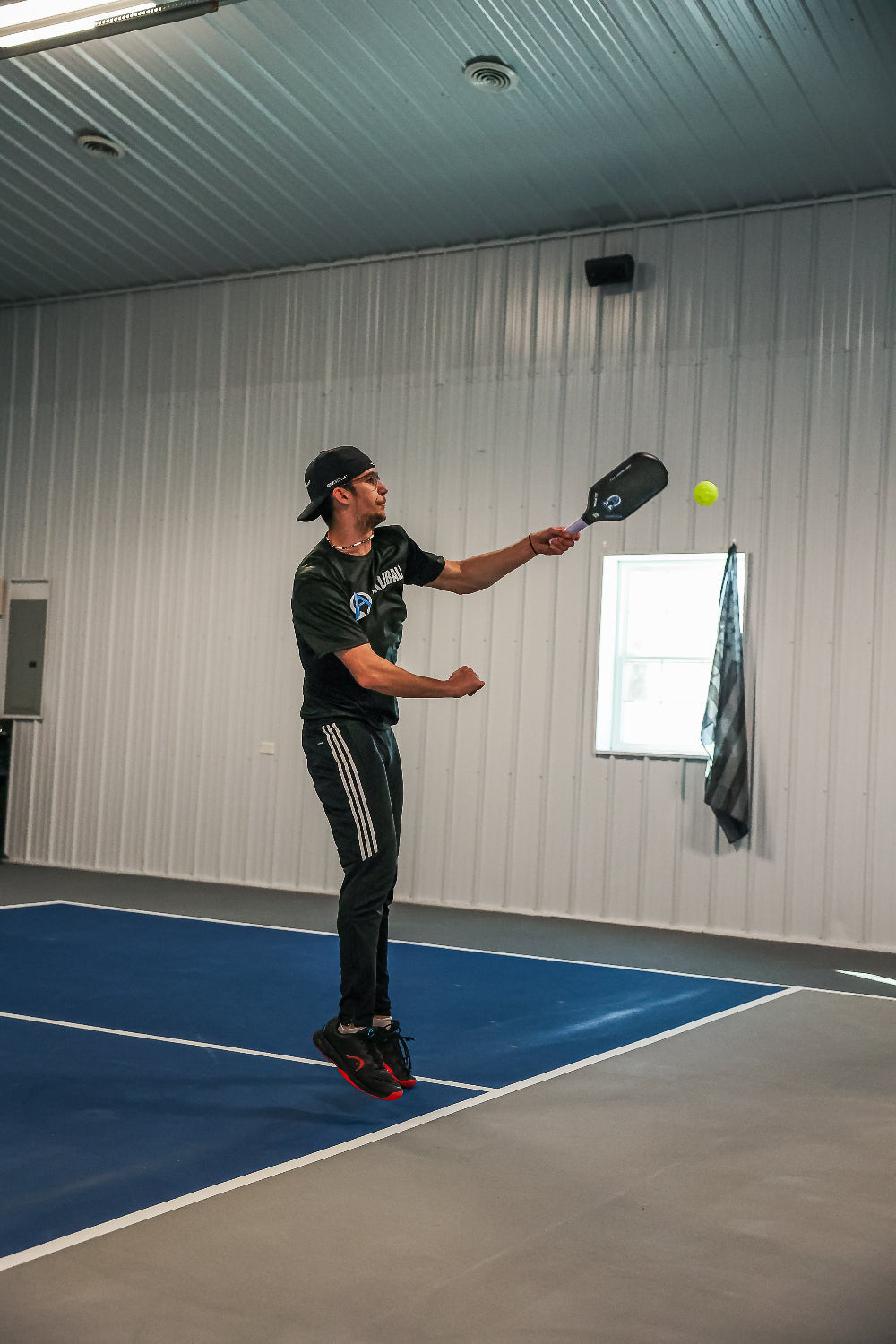 Man playing pickleball indoors, wearing black athletic clothing, red-soled shoes, and a baseball cap.