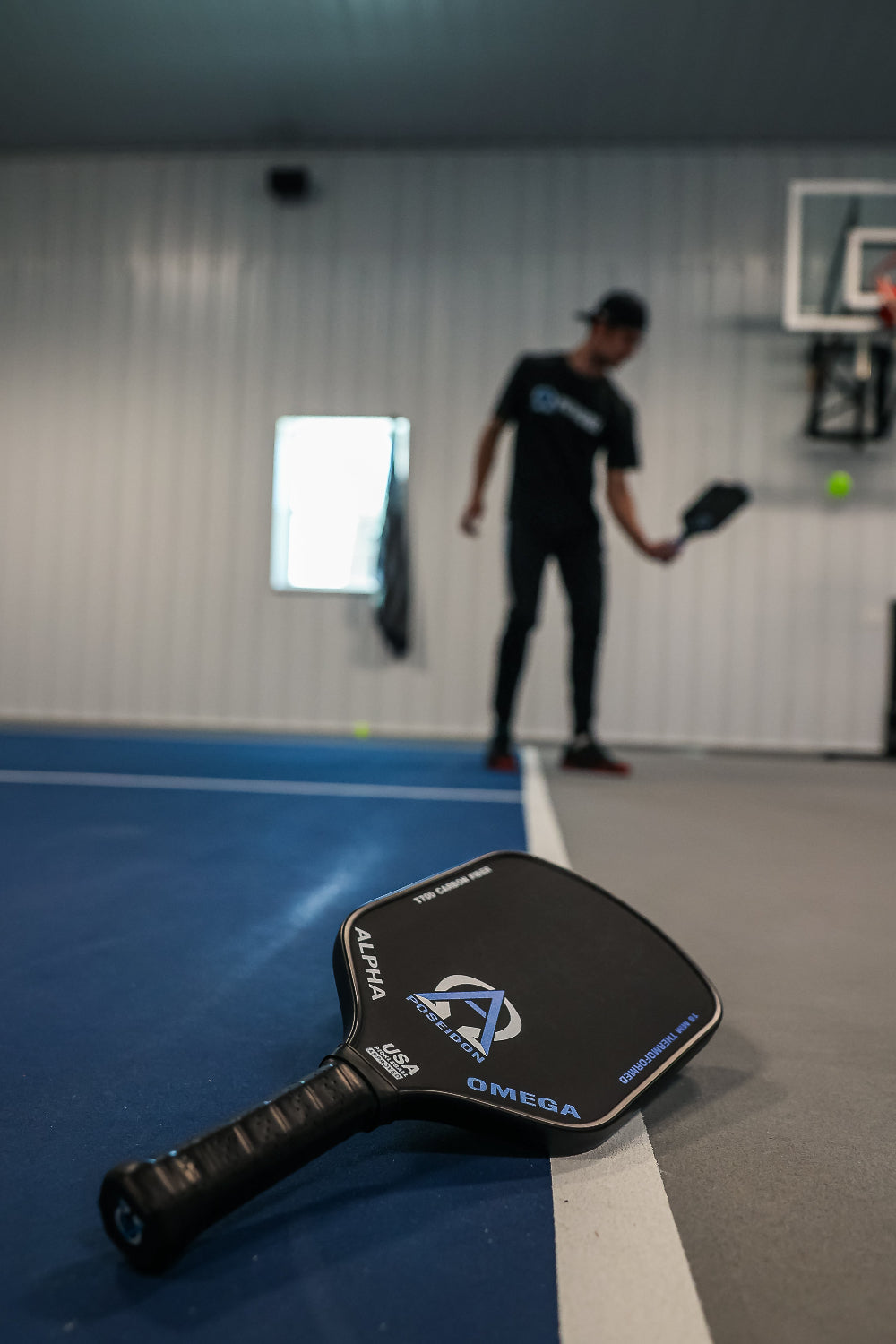 Close-up of a black Alpha Poseidon Omega pickleball paddle on a blue and gray court, with a blurred figure in a black shirt and pants holding another paddle in the background.