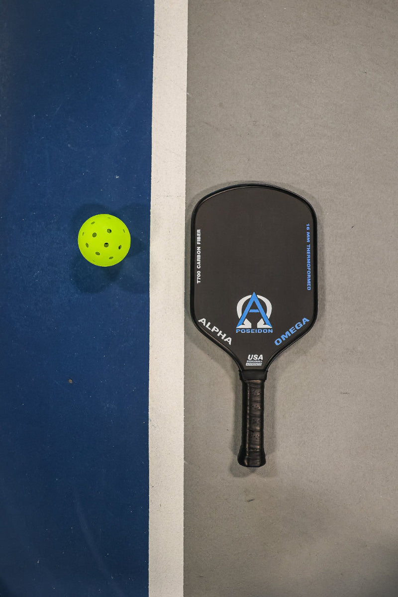 Overhead shot of a yellow pickleball, a black pickleball paddle, and a white line on a blue and gray pickleball court.