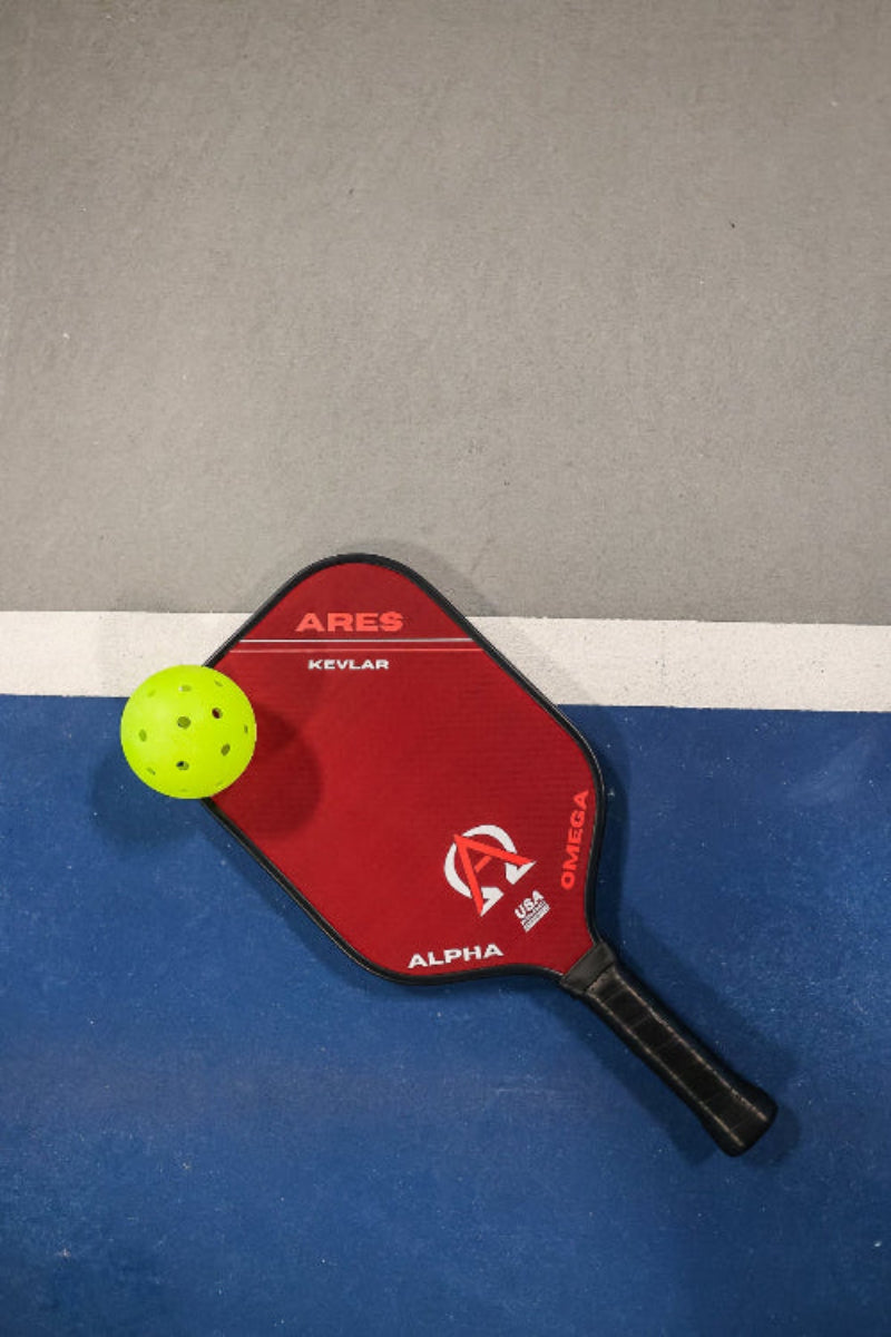 Red pickleball paddle with yellow pickleball on a blue and grey court.