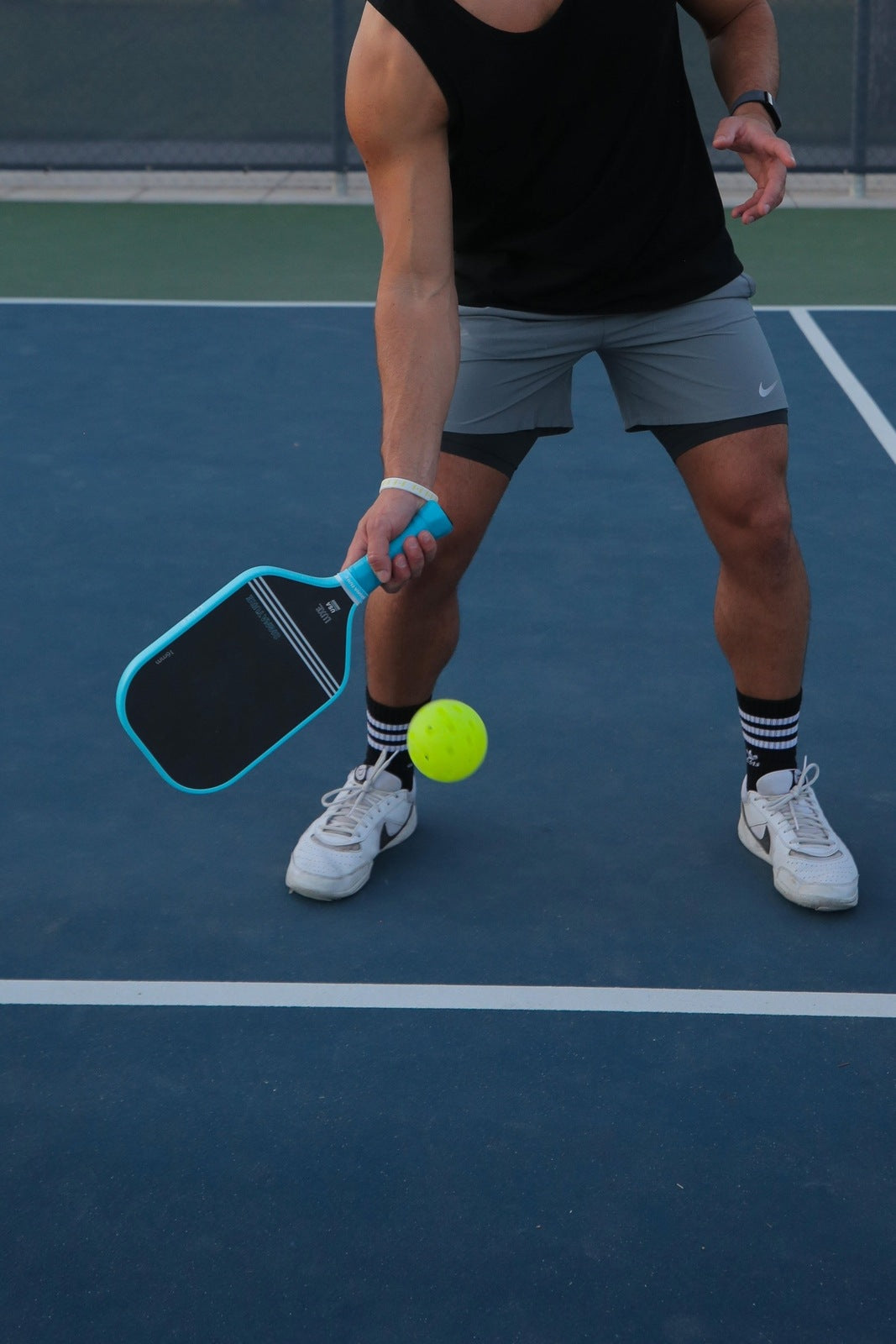 Person playing pickleball on a blue court, wearing black shirt, grey shorts, white shoes, black and white socks.