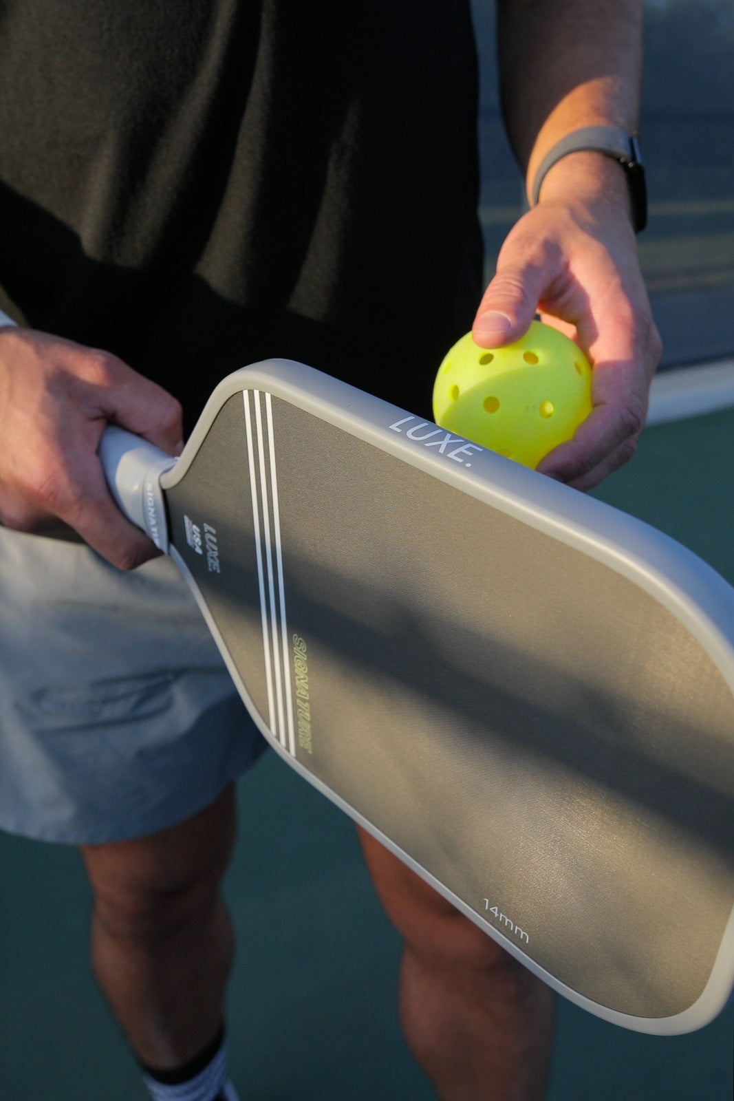 A person holding a pickleball paddle and a yellow pickleball ball. They are wearing a black shirt, grey shorts, and an Apple watch.