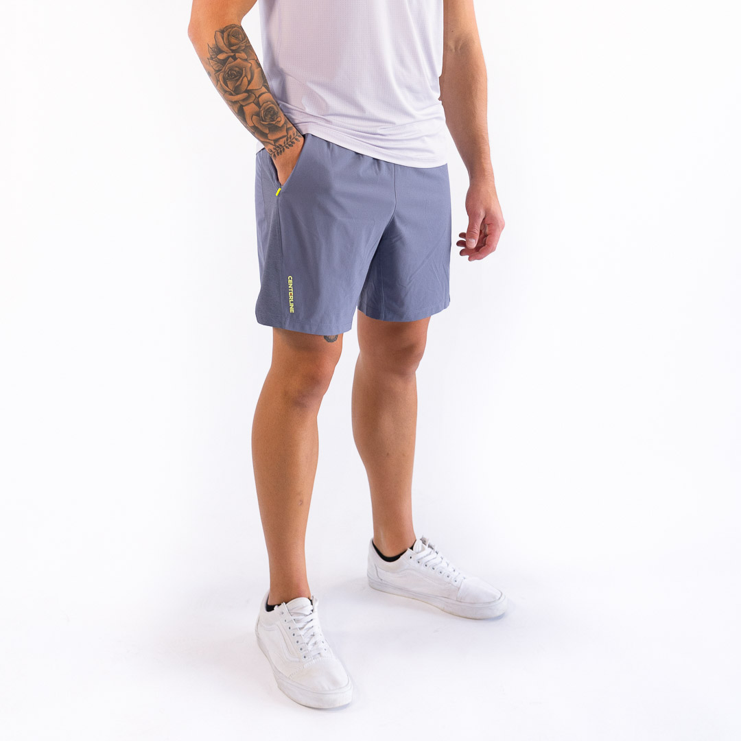 Man in white tank top and slate blue shorts with white sneakers. He has his hand in his pocket and a tattoo on his arm.
