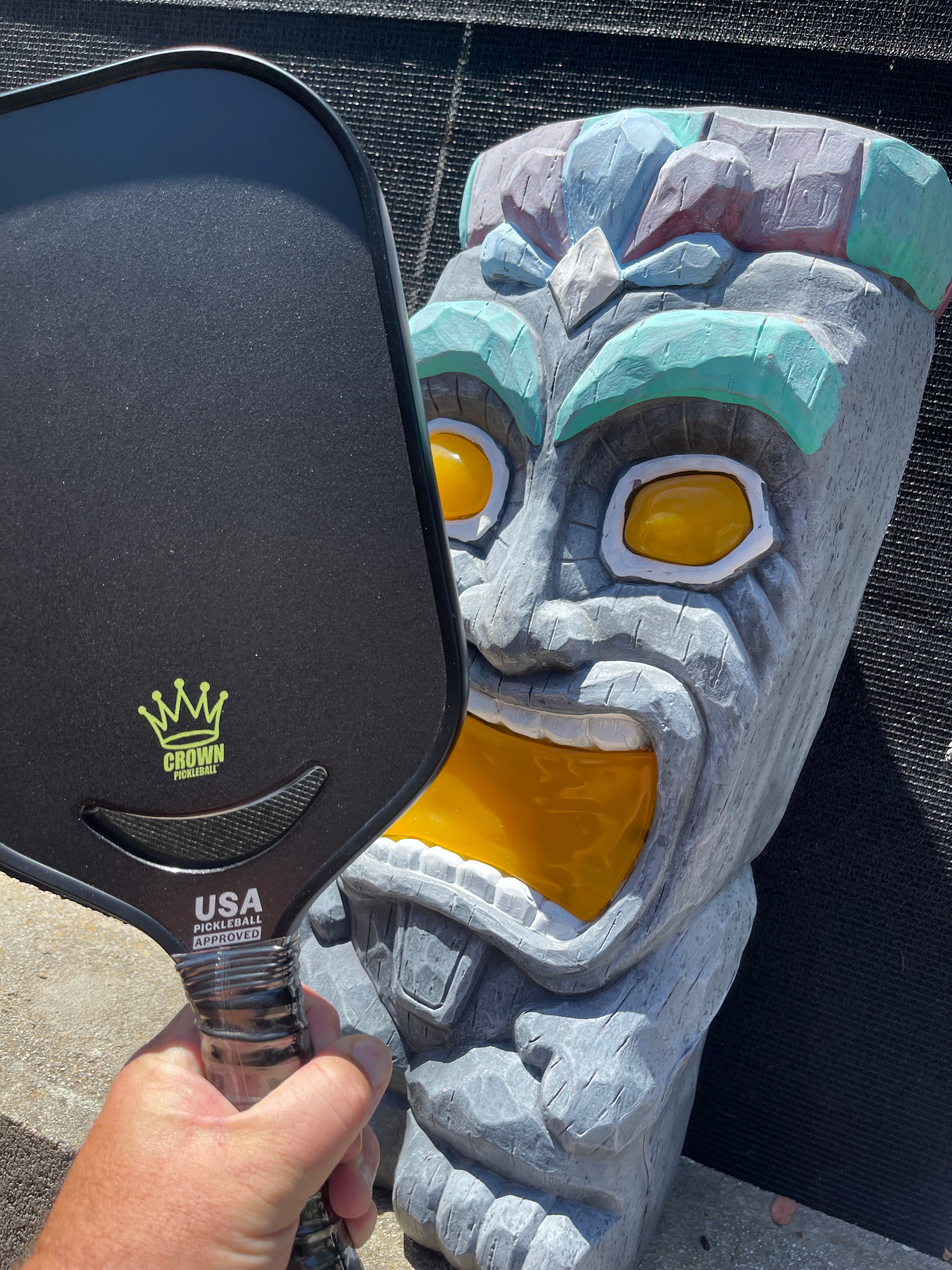 Hand holding a black Crown pickleball paddle next to a gray and colorful wooden tiki mask.