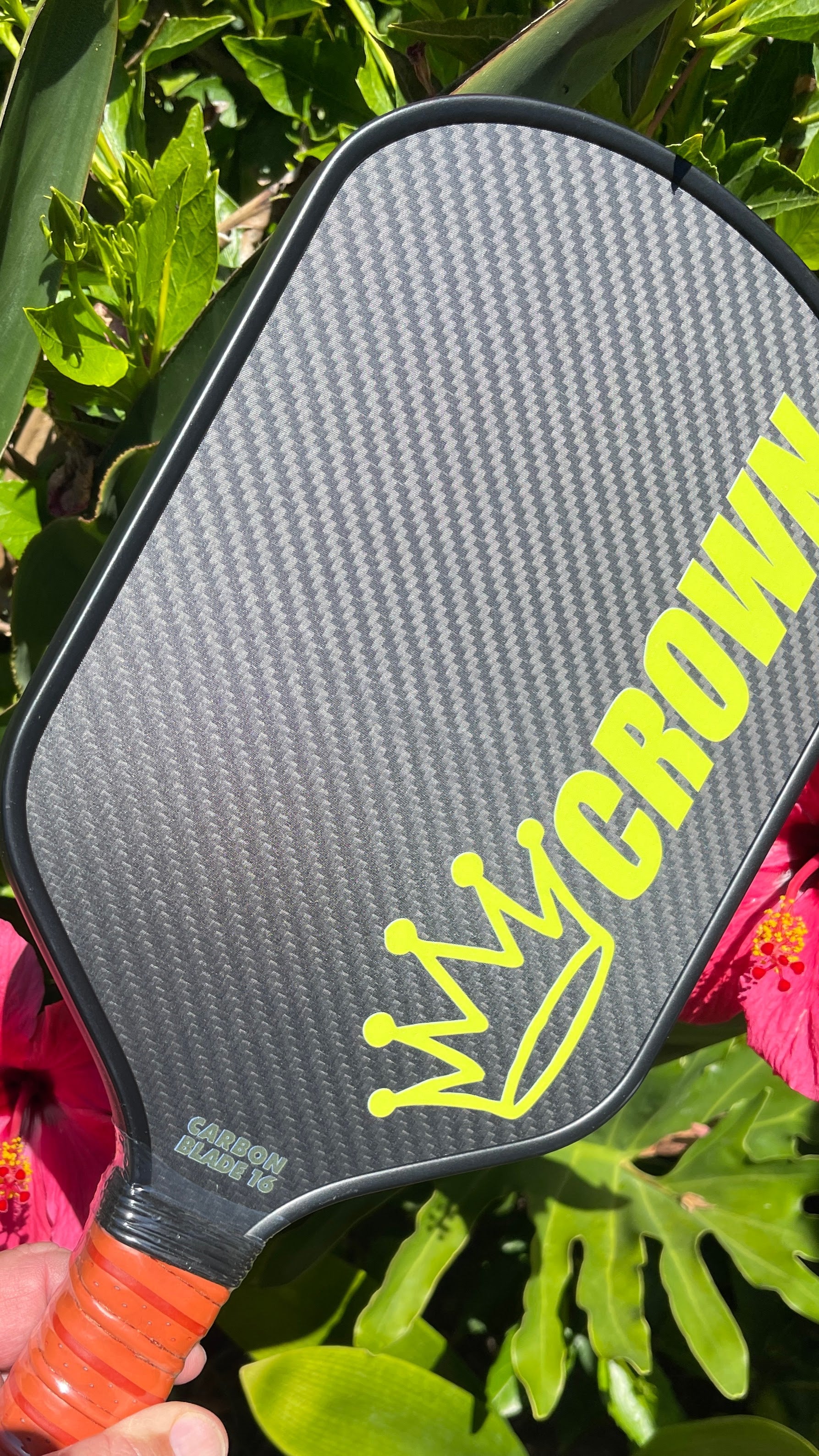 Close up of a gray and black Crown pickleball paddle with a yellow crown logo and the word "CROWN", held in front of green leaves and a pink hibiscus flower. 
