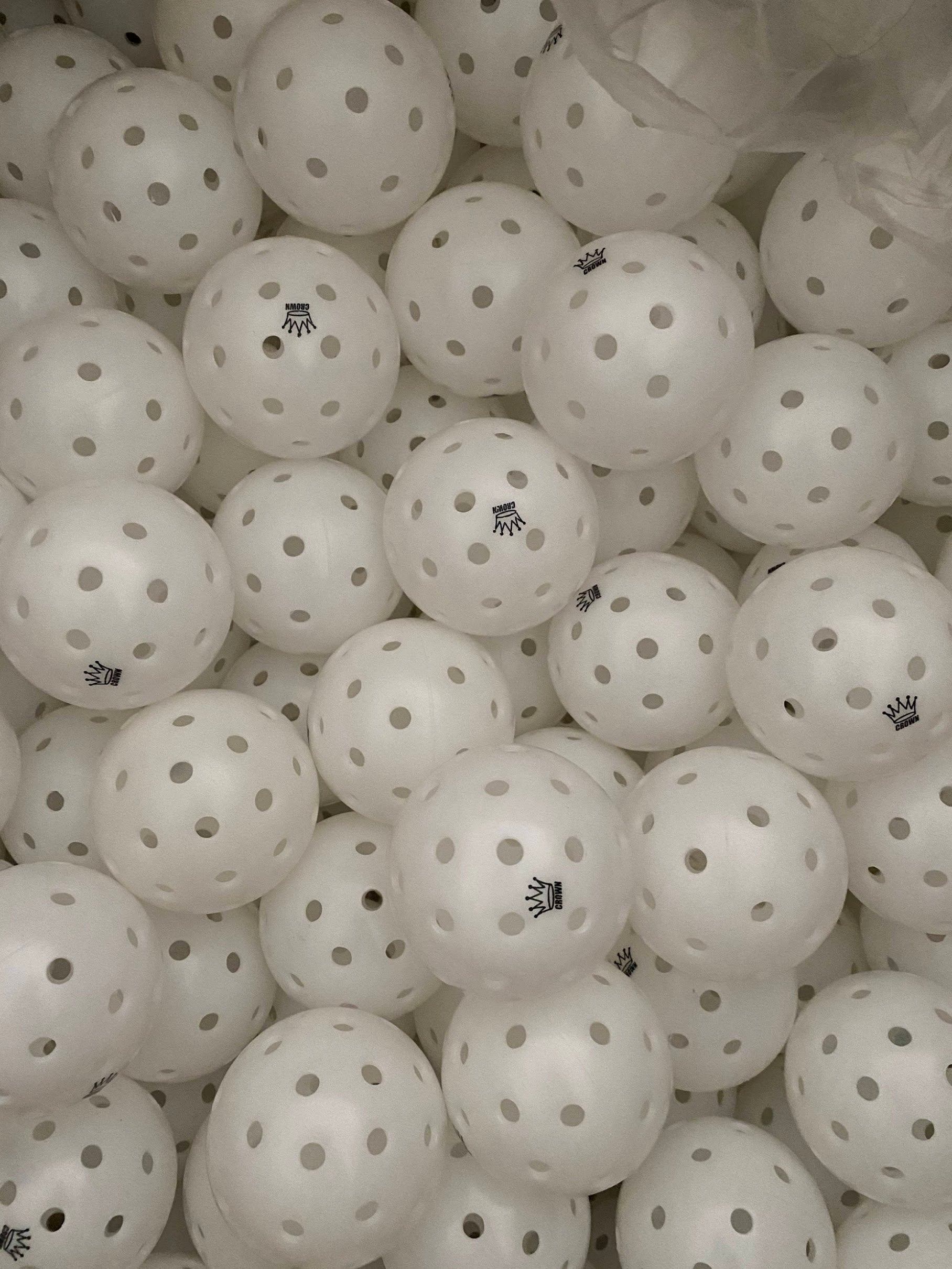 Pile of white perforated pickleballs with crown logos.
