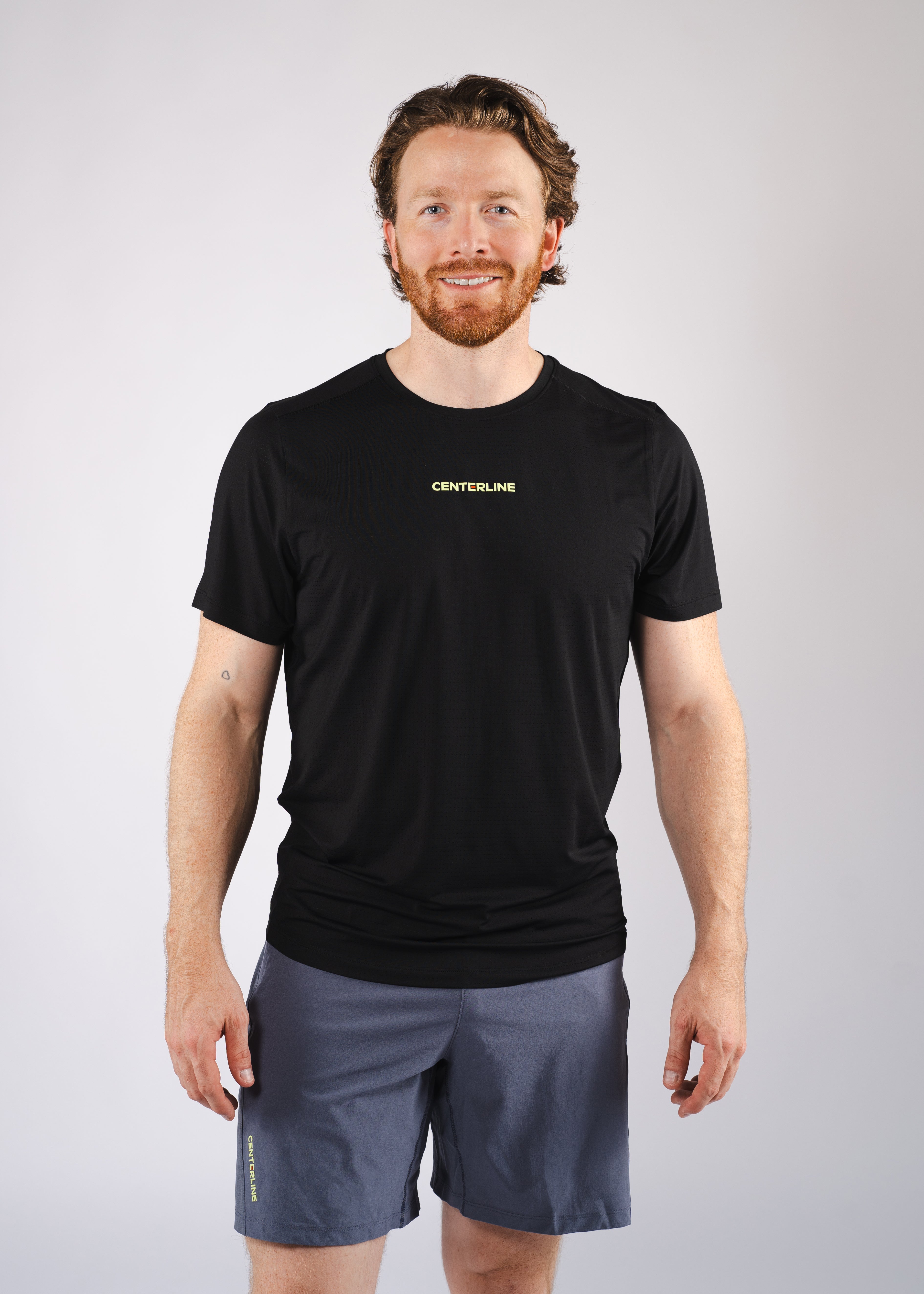A man in a black t-shirt and gray shorts stands smiling.
