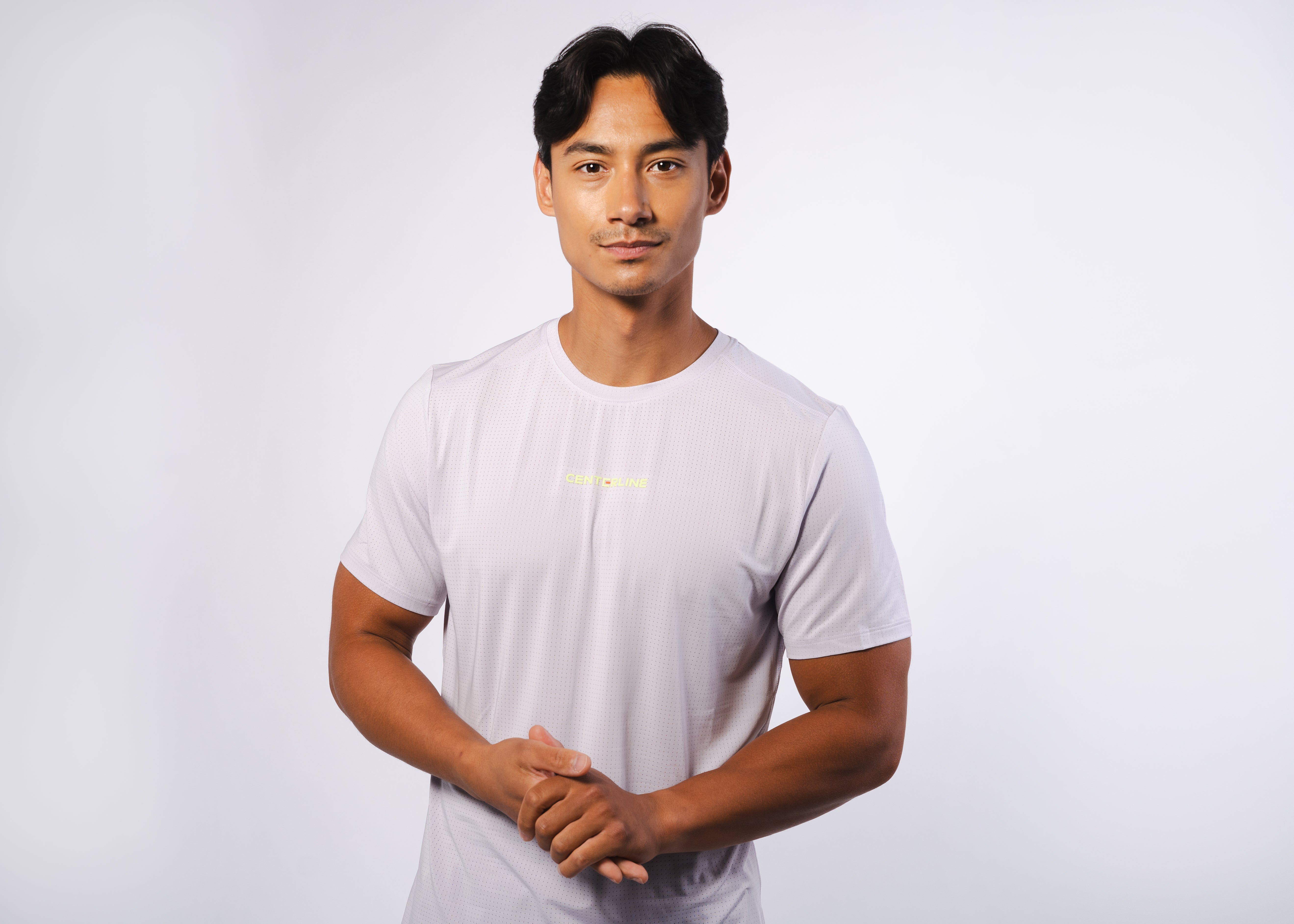 A man in a light purple athletic shirt with a subtle geometric pattern, hands clasped in front of him.