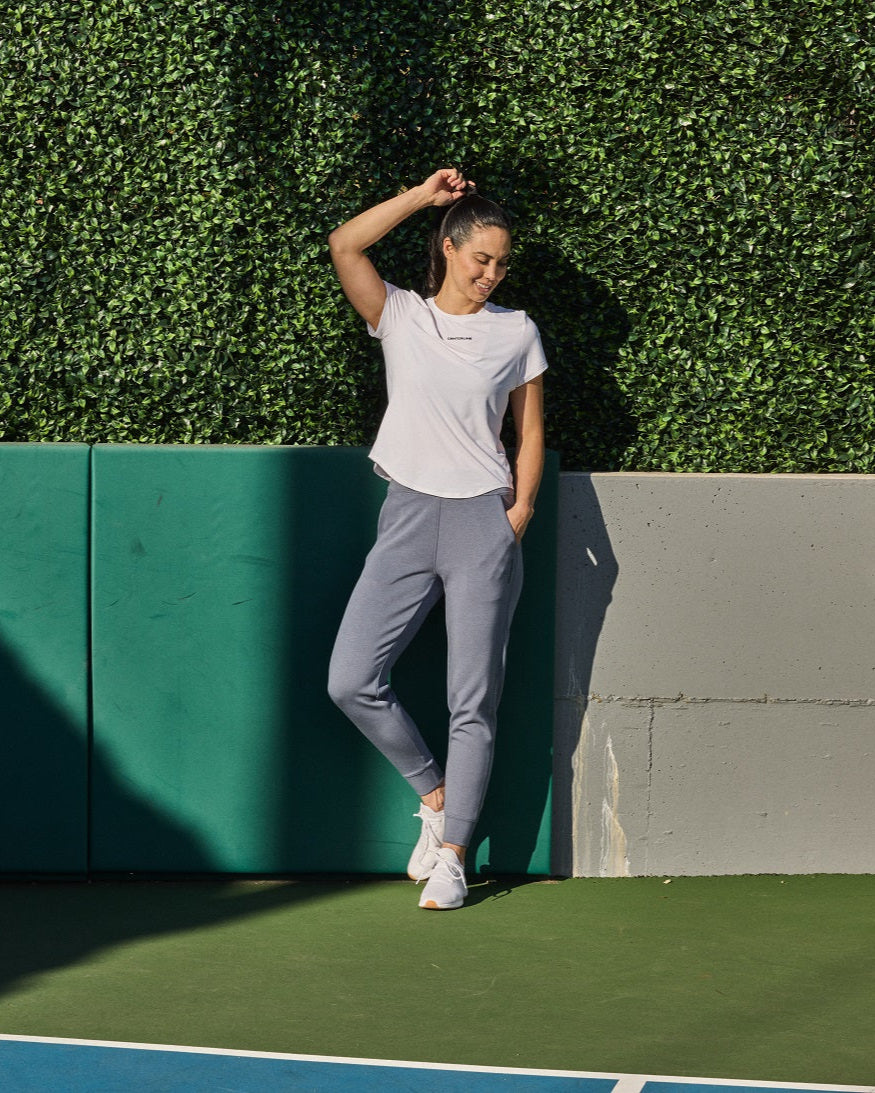 A woman in a white t-shirt and gray sweatpants stands smiling in front of a green hedge.
